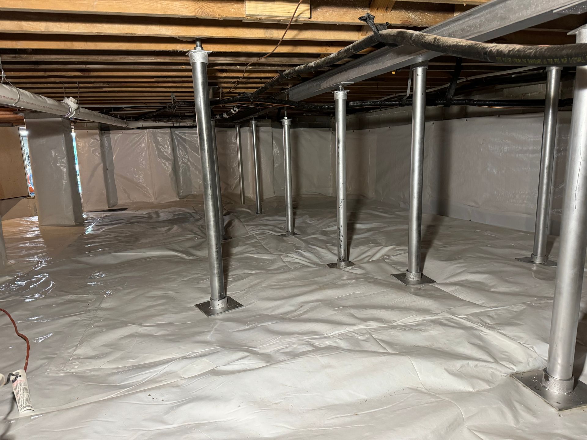 Crawl space with vapor barrier, supports, and metal beams under a house.