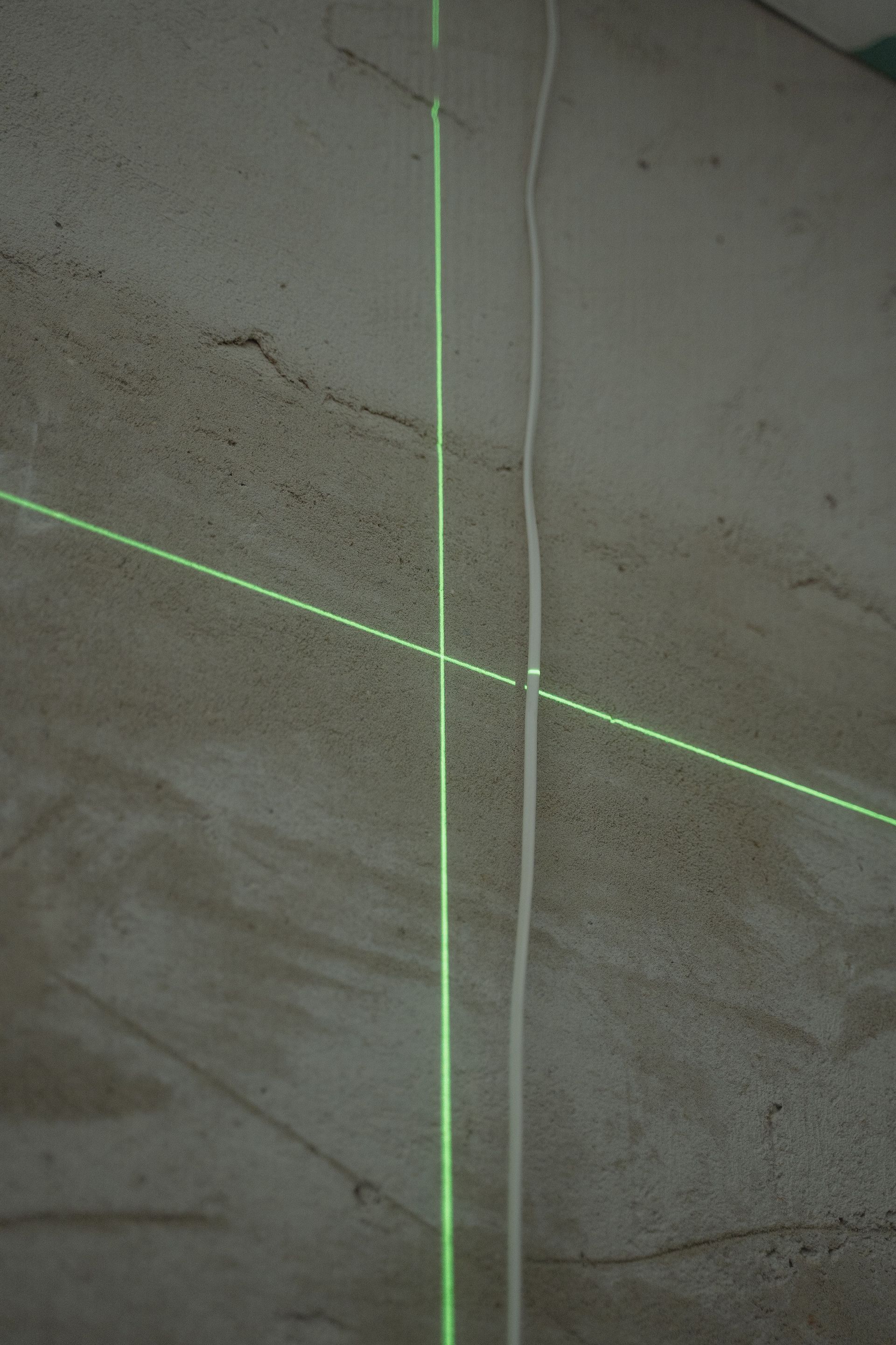 Green laser cross on a gray concrete wall with a white cable running vertically nearby.