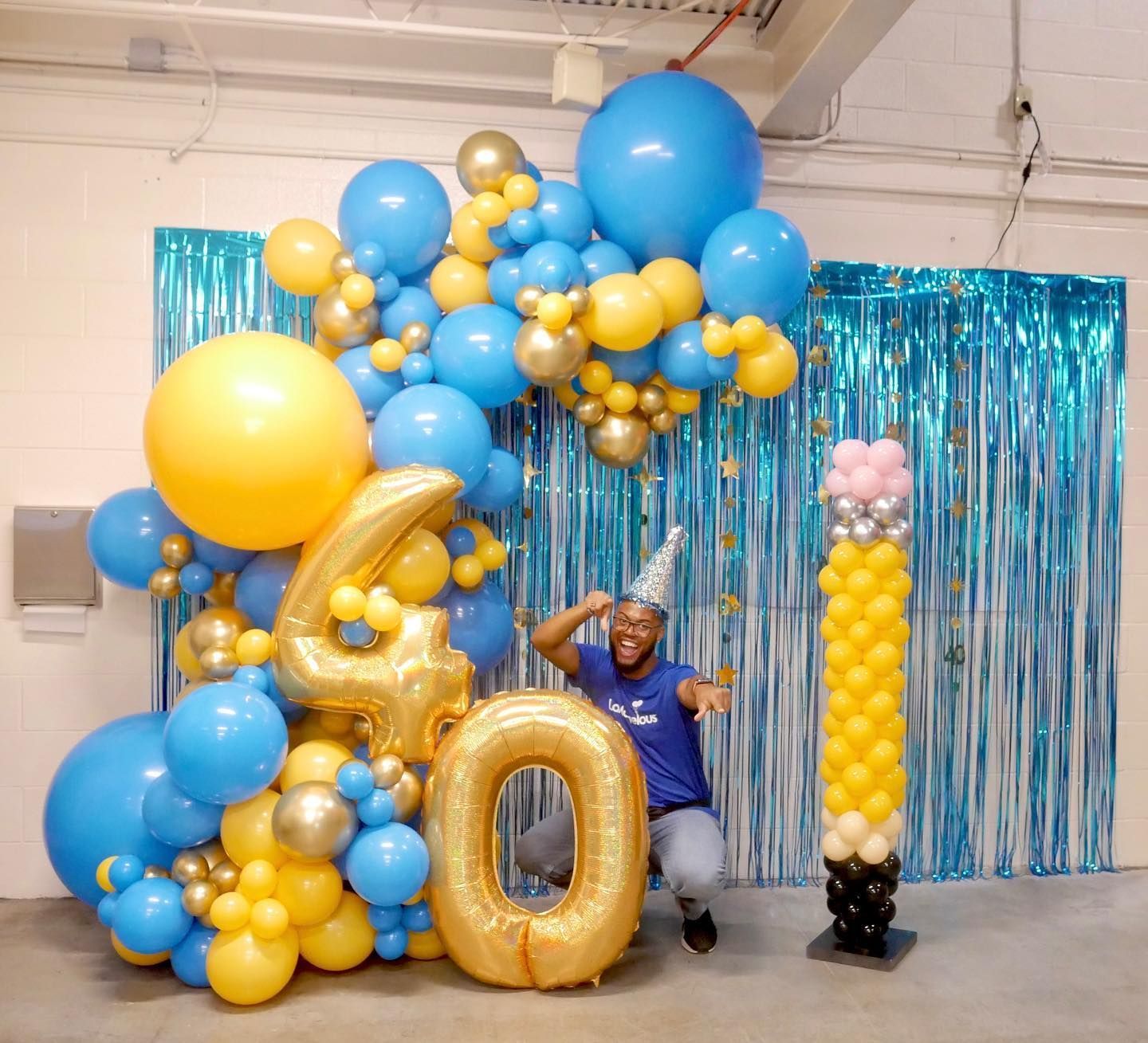 A man is standing in front of a wall covered in blue and yellow balloons.