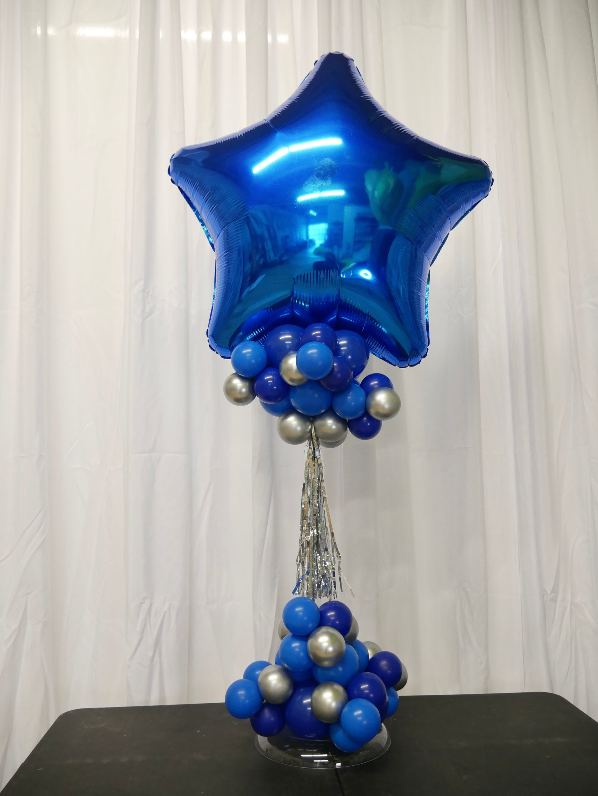 A blue star balloon is surrounded by blue and silver balloons on a table.