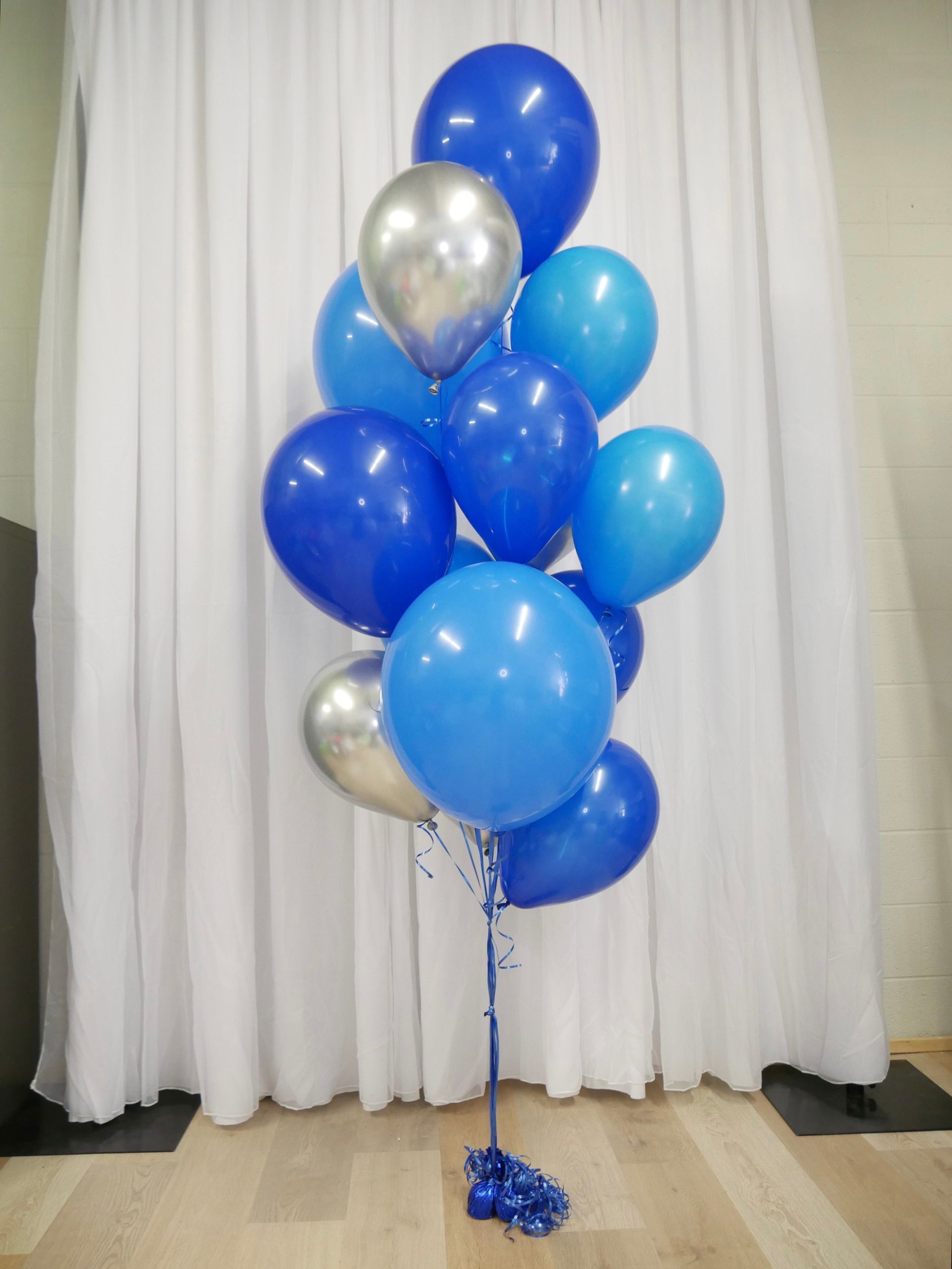 A bunch of blue and silver balloons are sitting on a wooden floor in front of a white curtain.