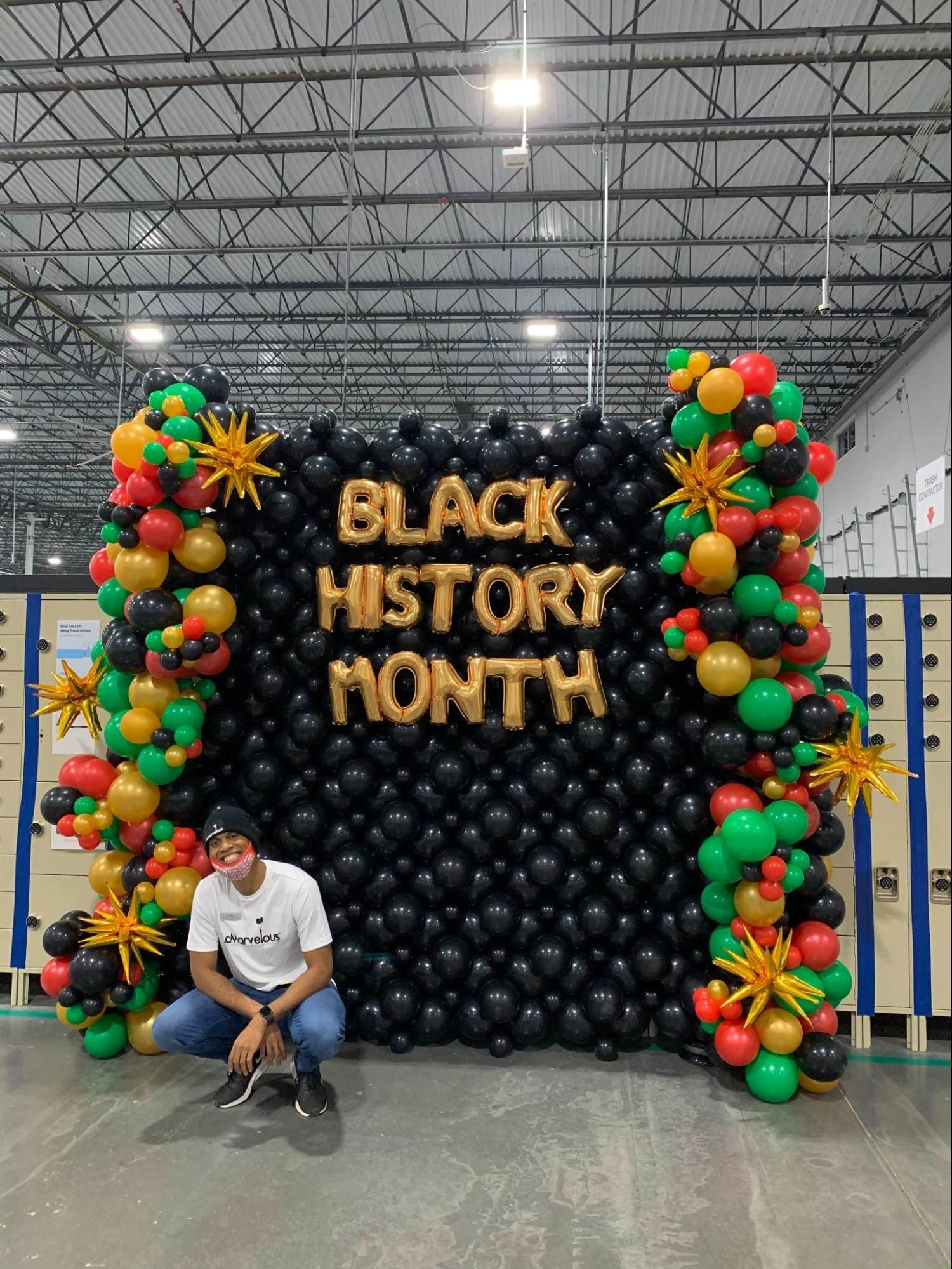 A man is kneeling in front of a wall made of balloons for black history month.