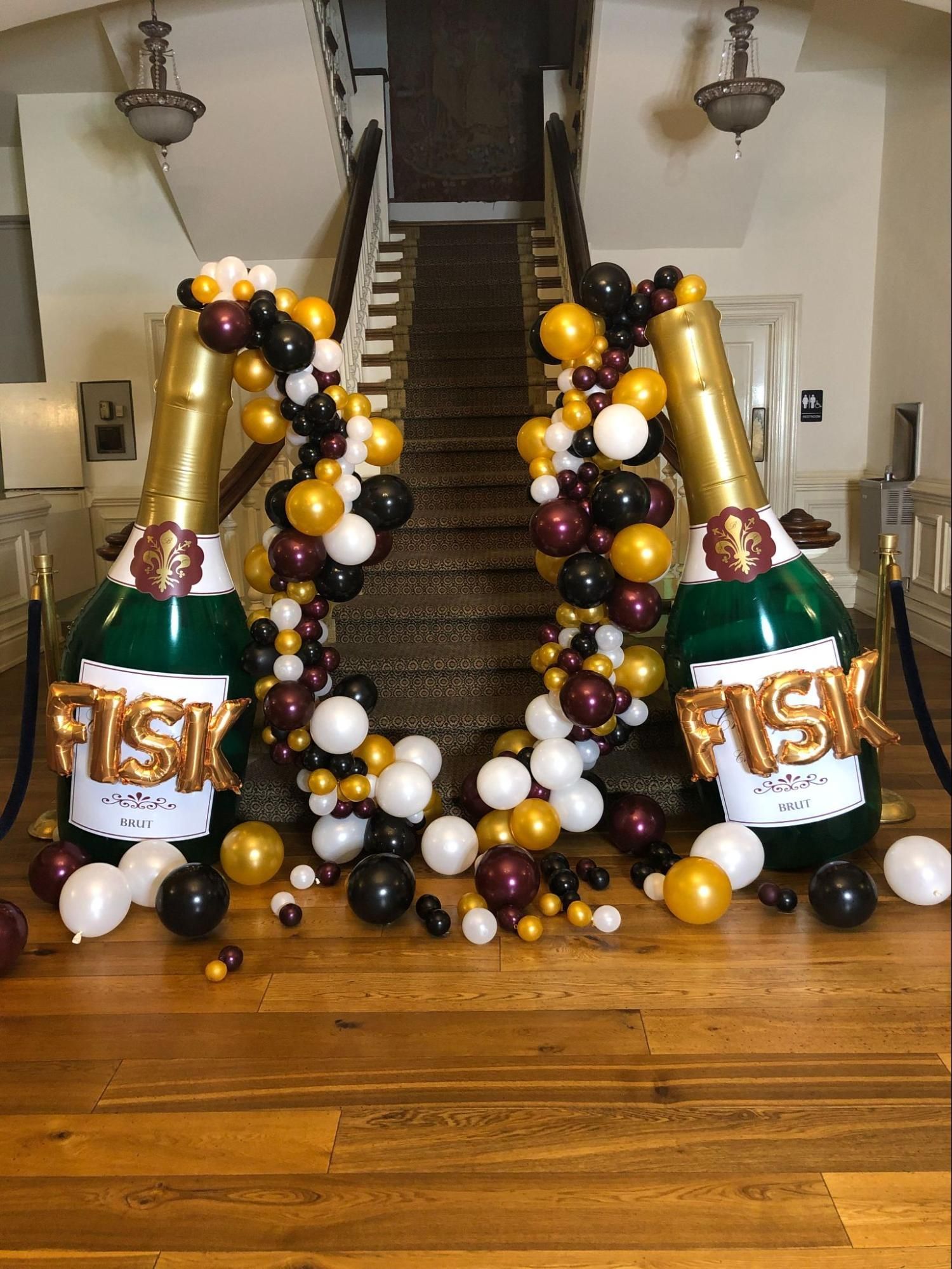 Two bottles of champagne made out of balloons are sitting on a wooden floor in front of a staircase.