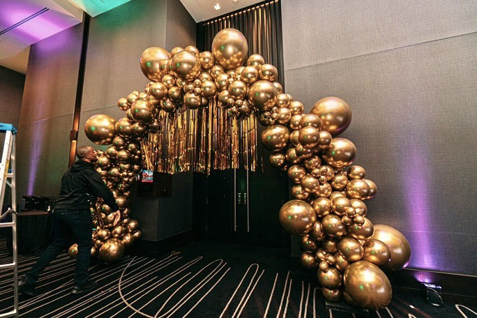 A man is standing in front of a large arch made of gold balloons.