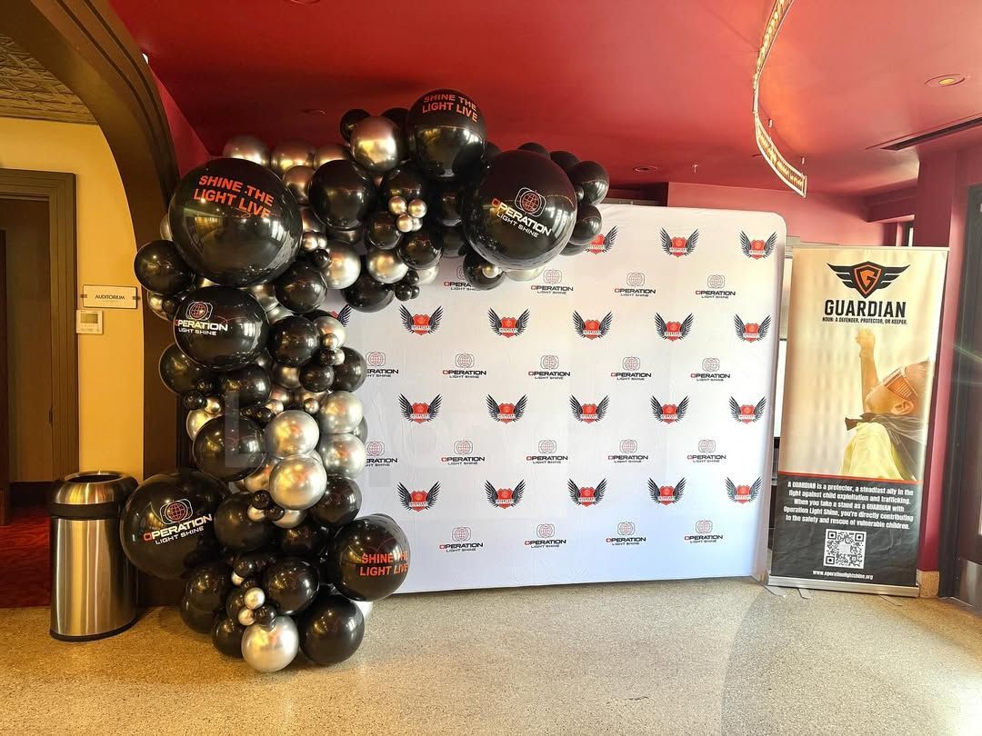 Black and silver balloon arch next to a white backdrop with logos and a sign in a venue.