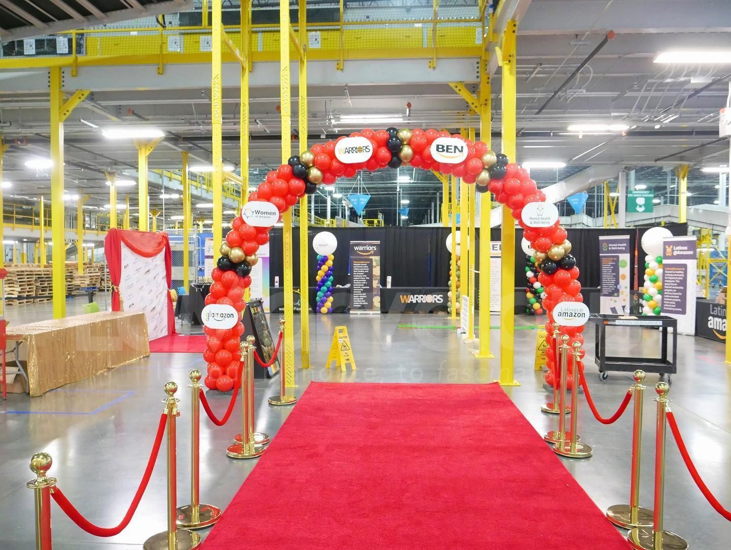 A red carpet is leading to a balloon arch in a warehouse.