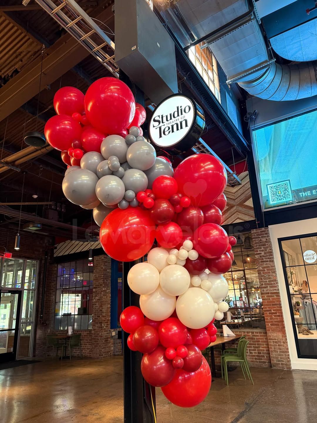 Red, gray, and white balloon arch decorates a black pole near a shop's entrance.