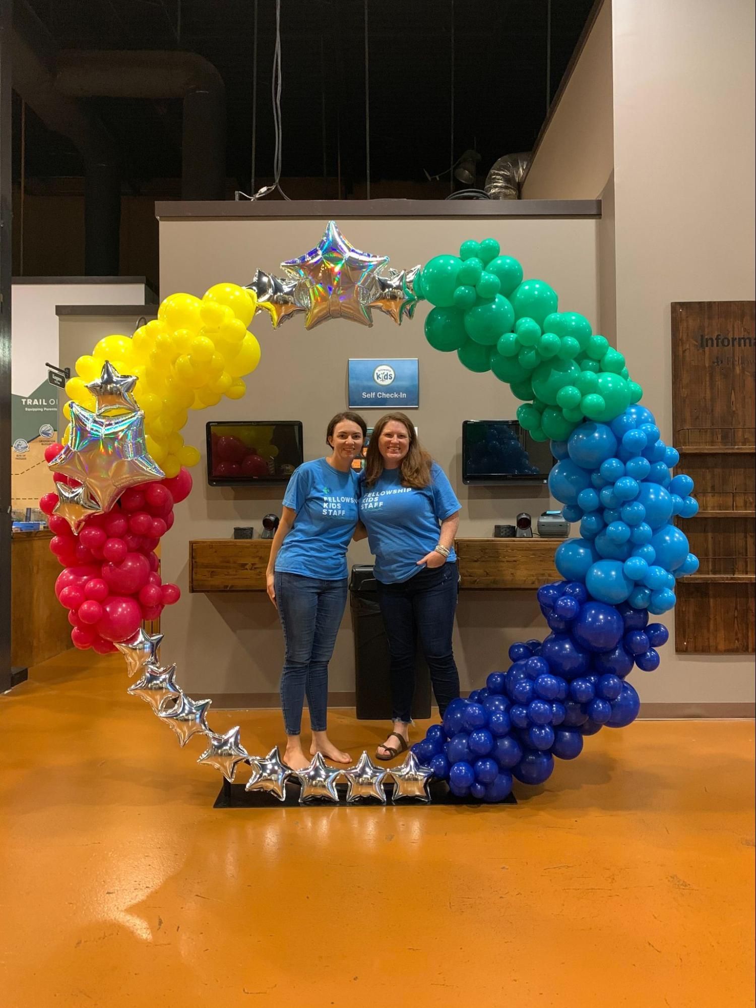 Two women are standing in front of a rainbow colored balloon arch.
