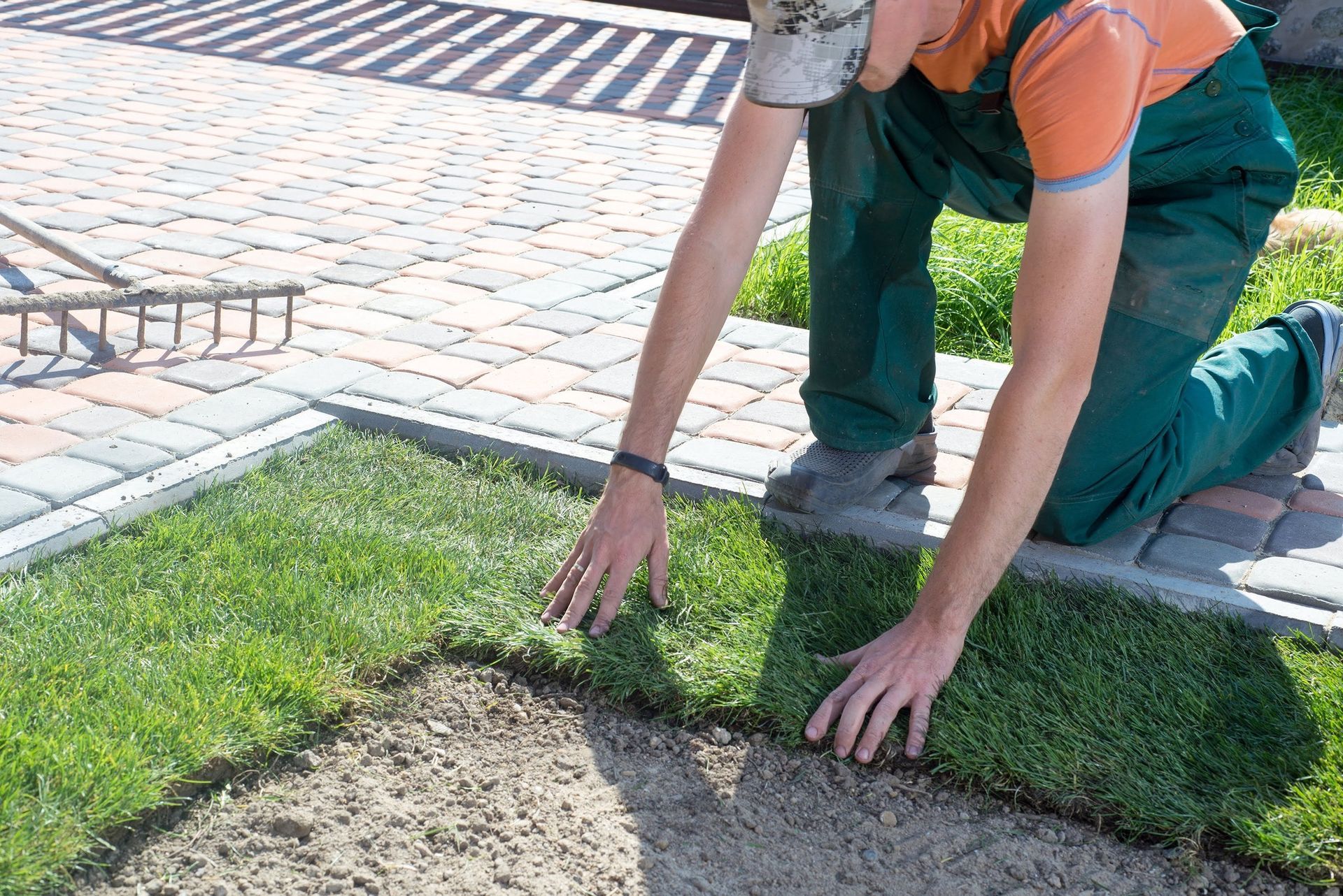 A man is kneeling on a brick sidewalk while raking grass.