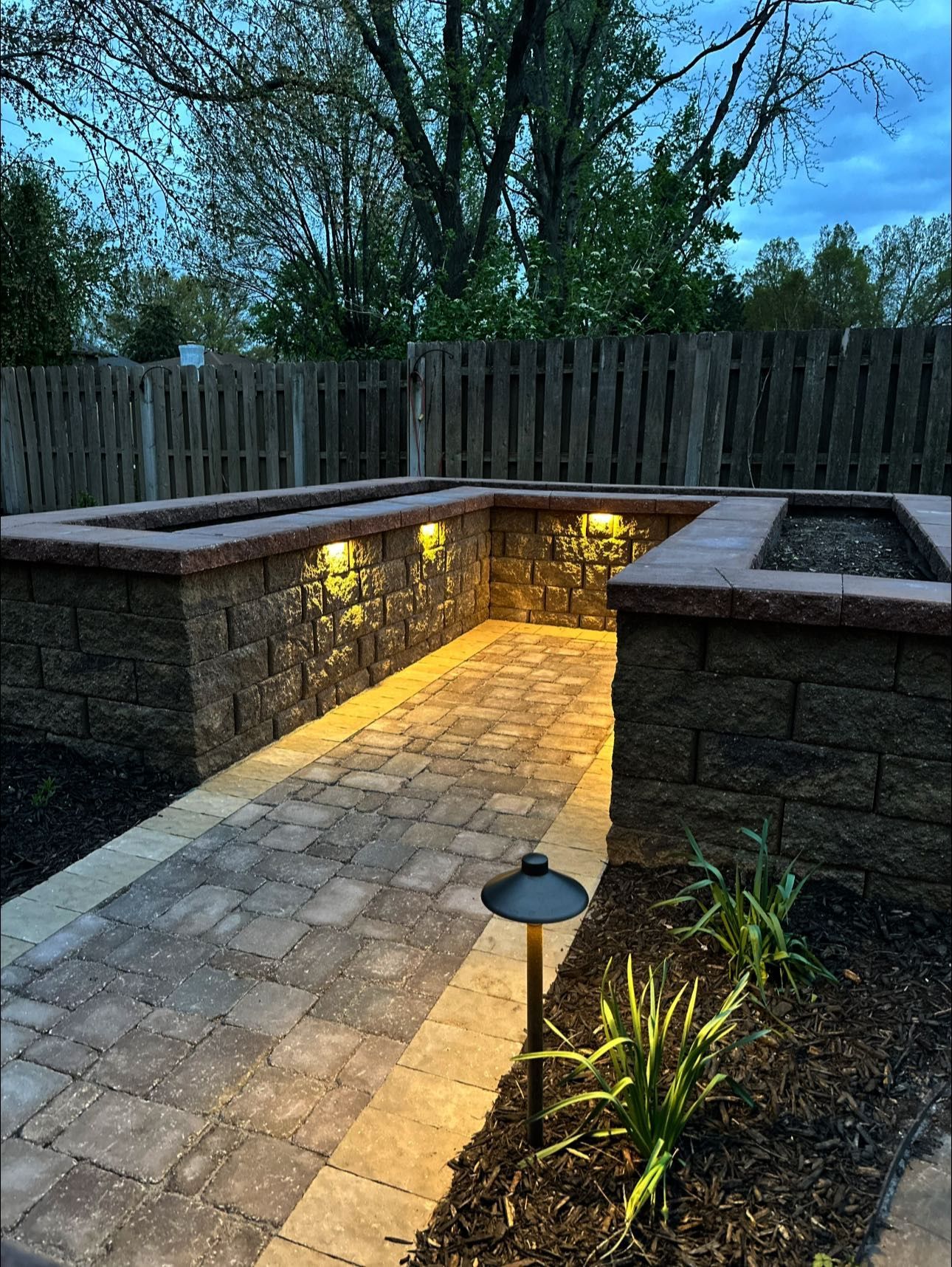 A brick walkway with lights on the side of it and a wooden fence in the background.