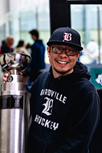 Smiling person in a black hoodie and cap posing beside a championship trophy