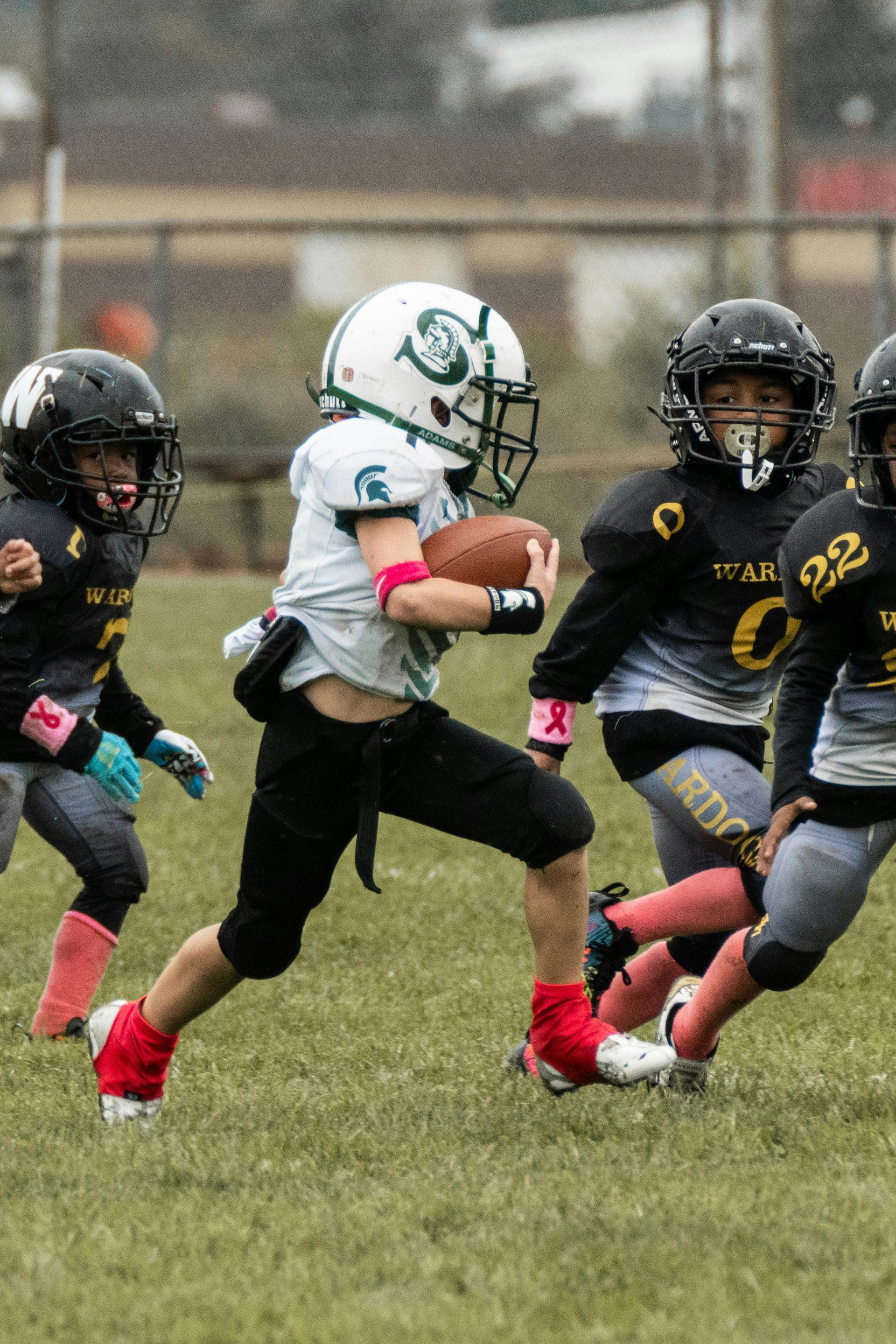 Youth football player in white runs with the ball while black-clad defenders close in on a grassy field