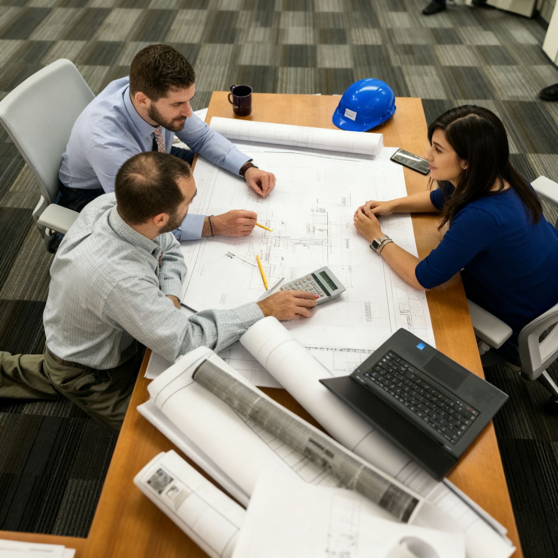 Three people collaborating around a table covered in blueprints and a laptop; one wears a blue hard hat.