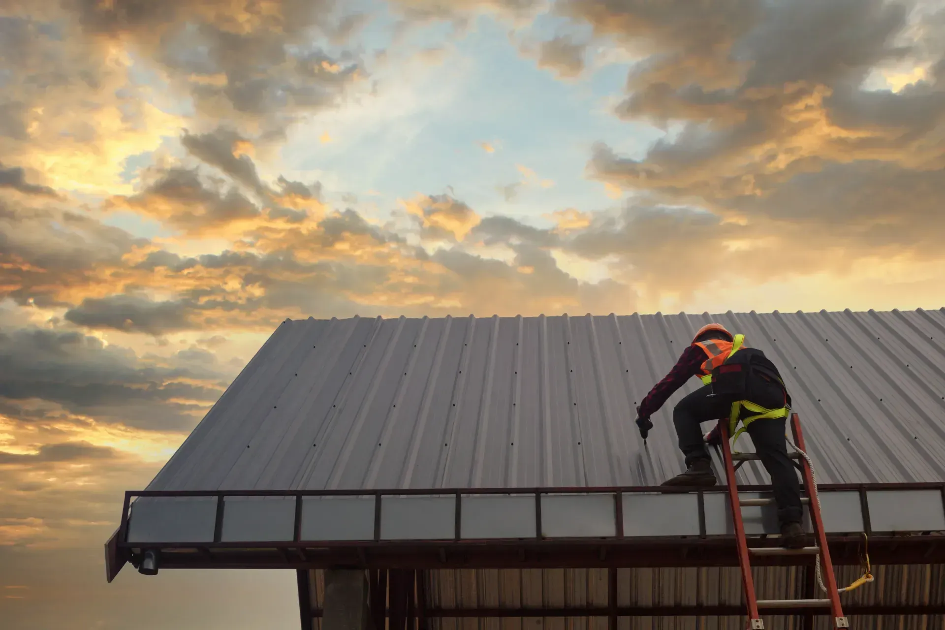 Roofer wearing safety gear on a metal roof, reaching down from a ladder; sunset sky.
