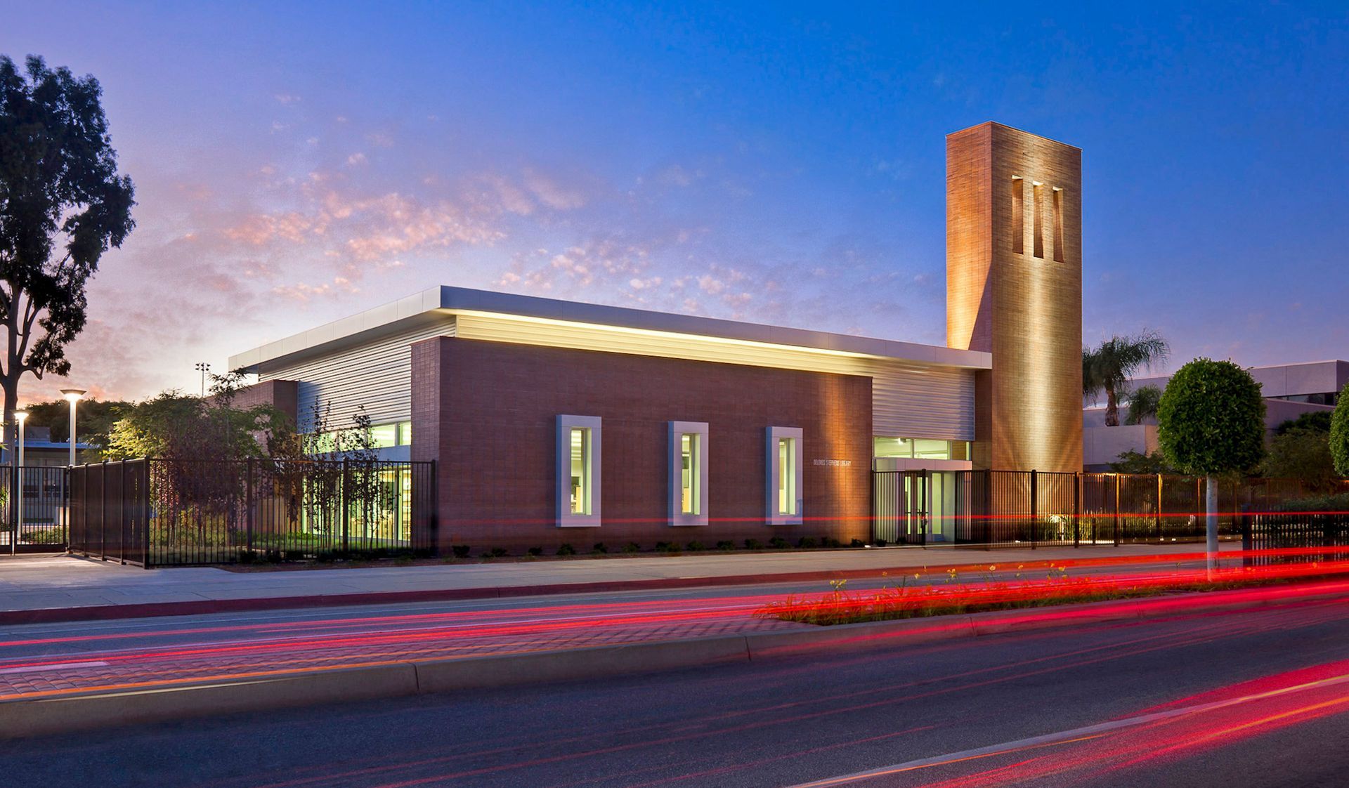 Brick building with a tower, lit at dusk. Red car lights streak across a street in the foreground.