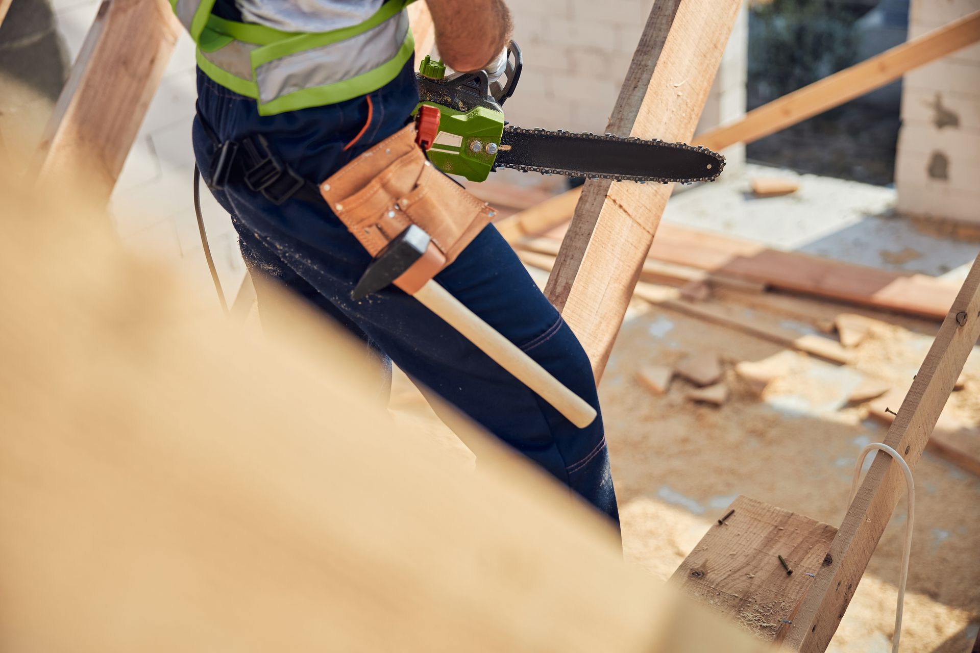 Carpenter using a chainsaw on a wooden structure, wearing a safety vest and carrying tools in a belt.