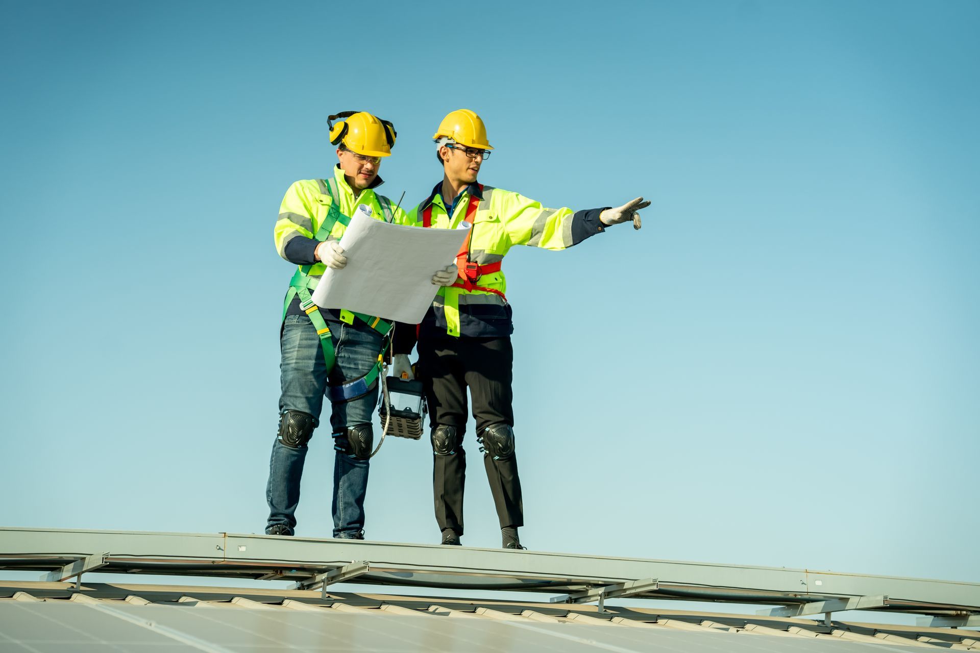 Two workers in safety vests and helmets on a rooftop, reviewing plans under a clear blue sky.