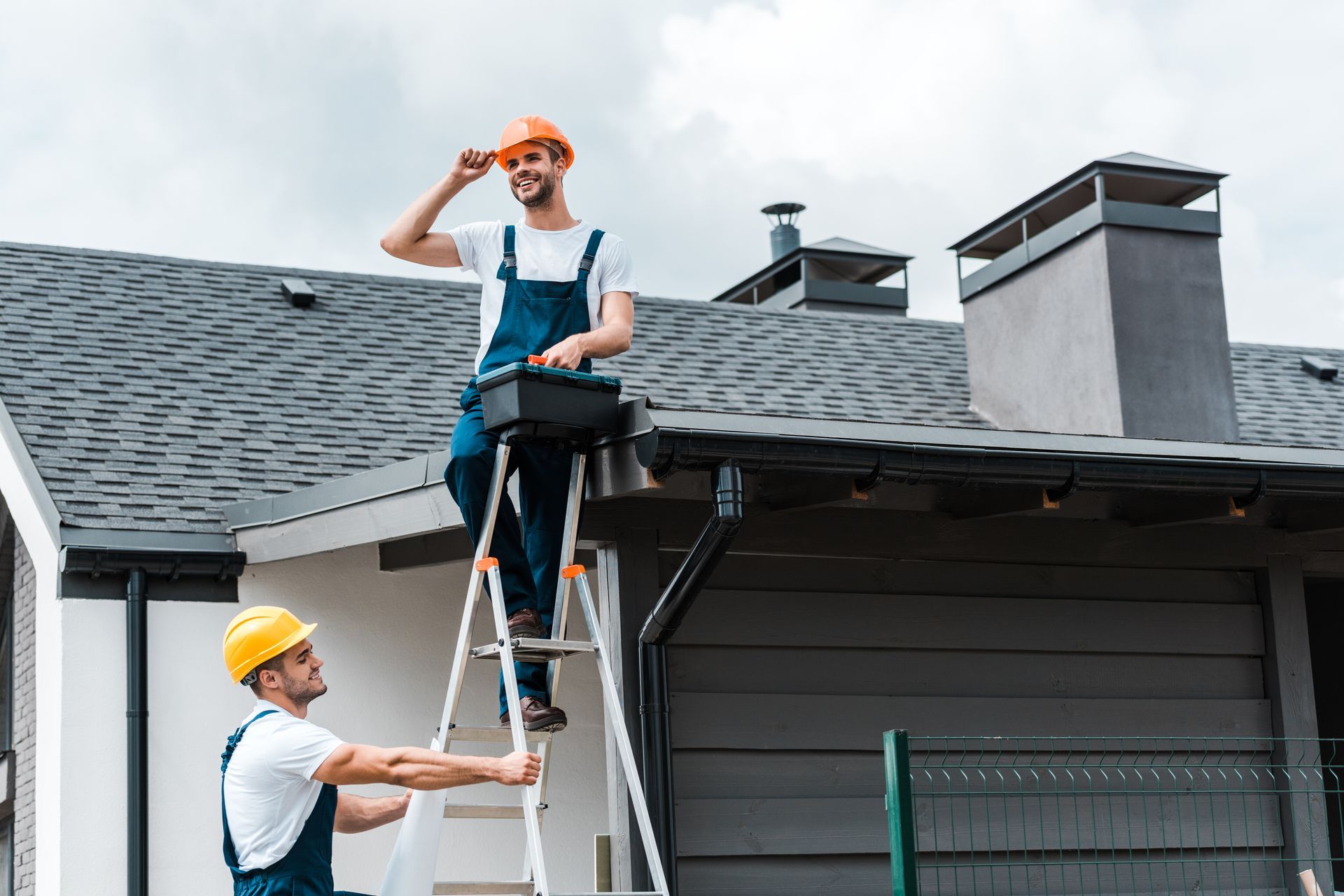 Two construction workers on a ladder, one adjusting a hard hat while the other steadies the ladder on a roof.