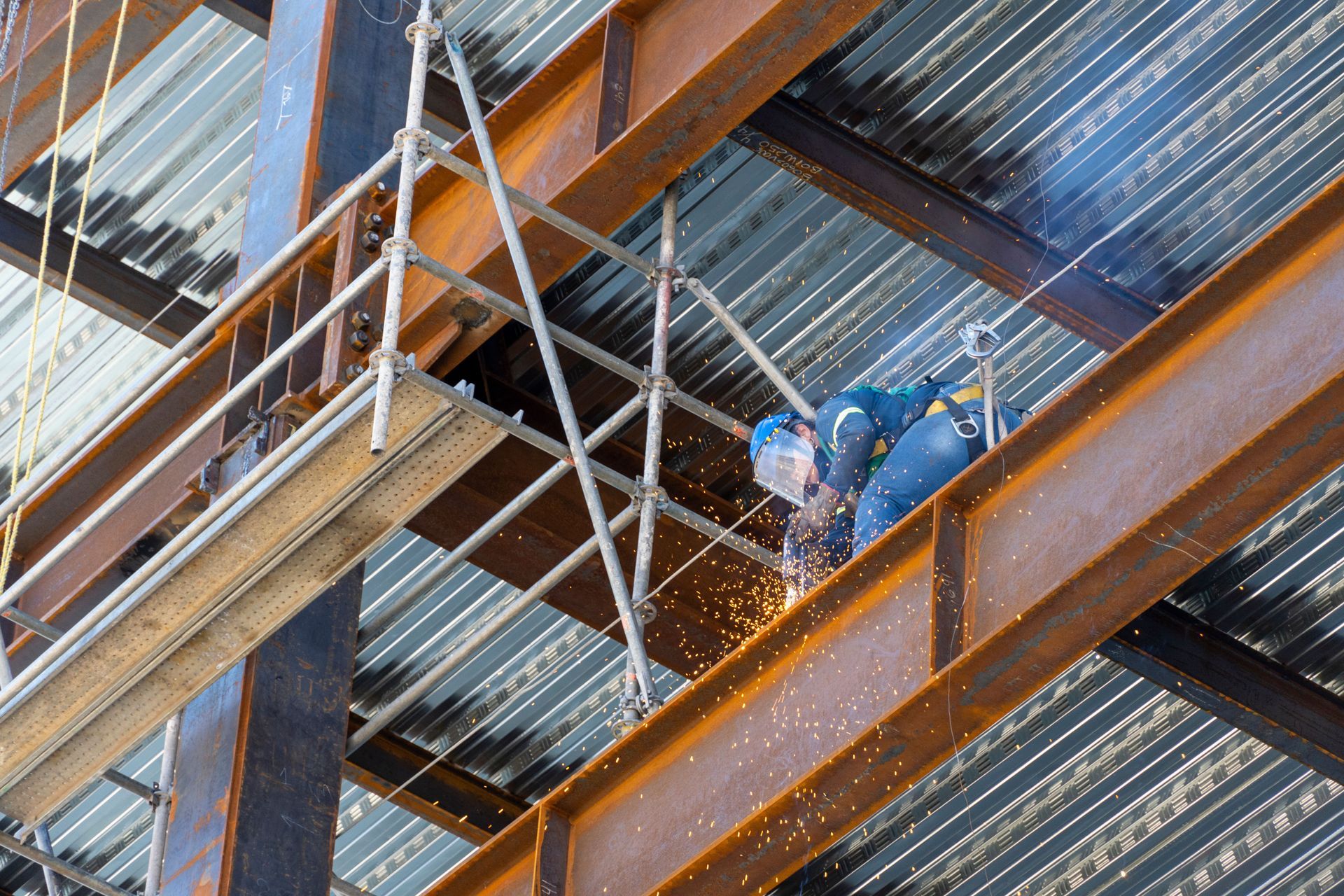 Construction worker welding steel beam on high-rise building with safety harness and scaffolding. Sparks fly.