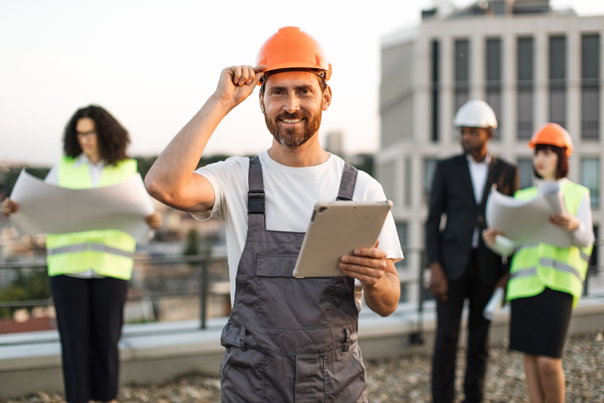 Construction worker with orange hard hat and tablet, smiling on a rooftop with colleagues.