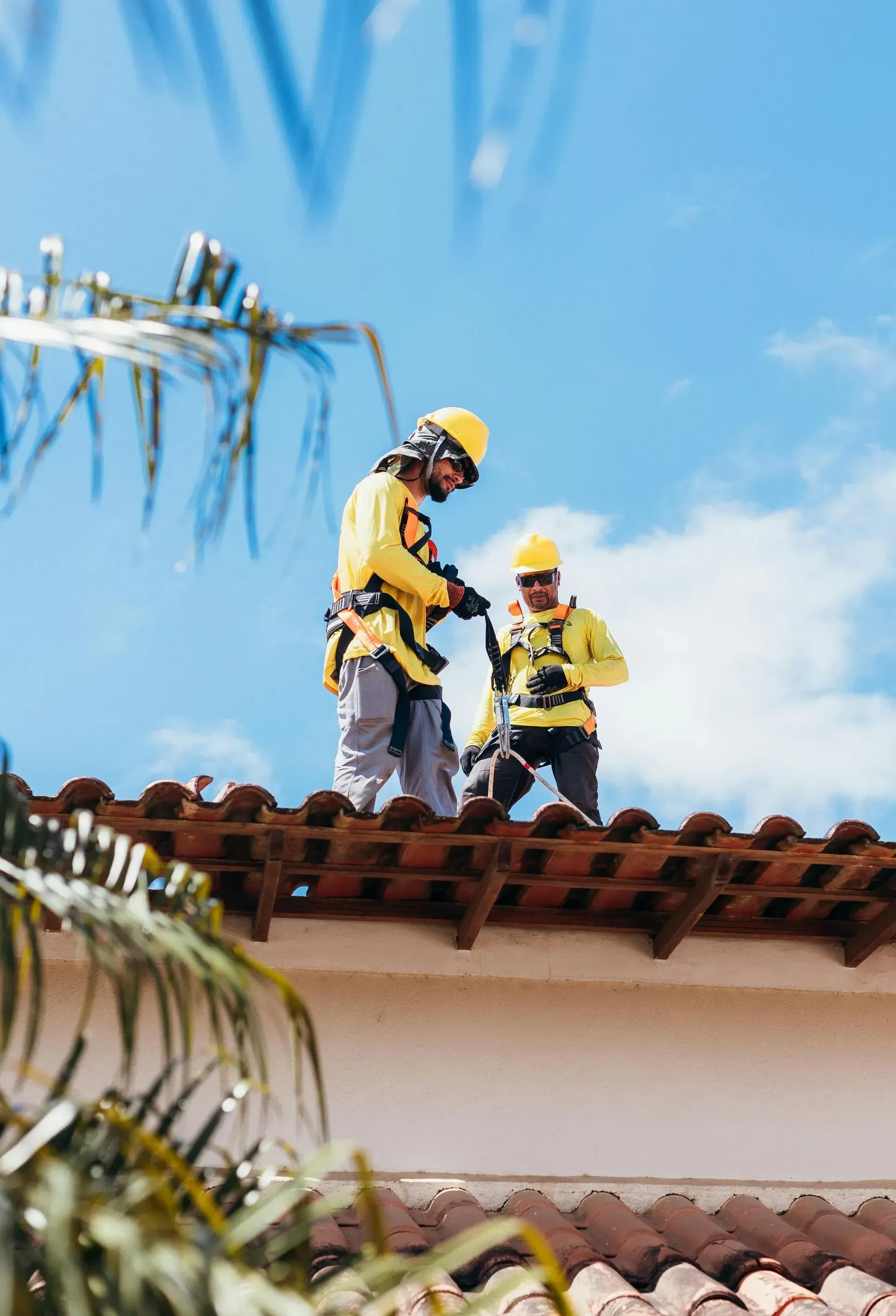 Two workers in safety gear on a tiled roof under a blue sky, one adjusting equipment.