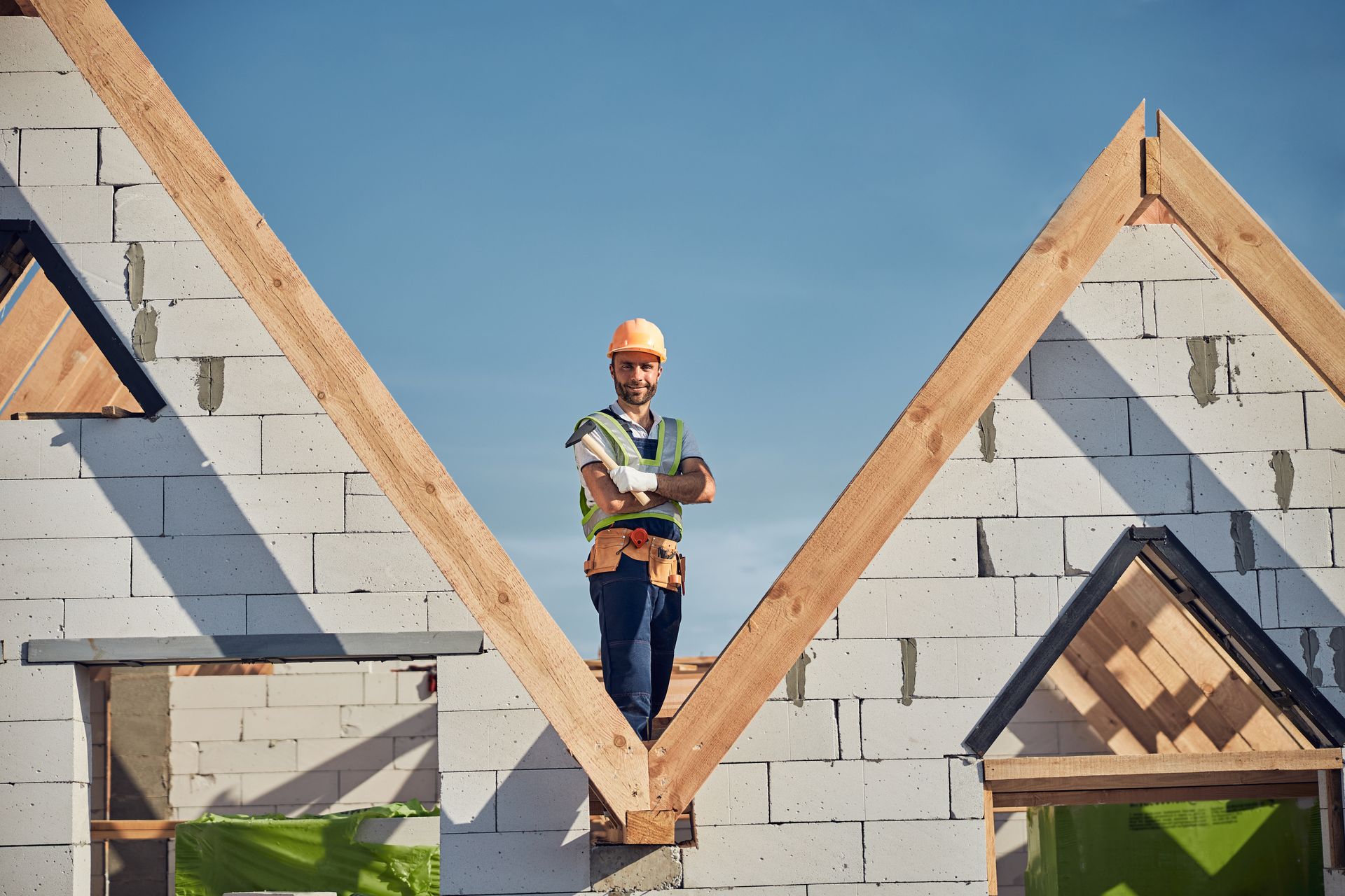 Construction worker stands on wooden beams, arms crossed, at a building site, against a clear blue sky.