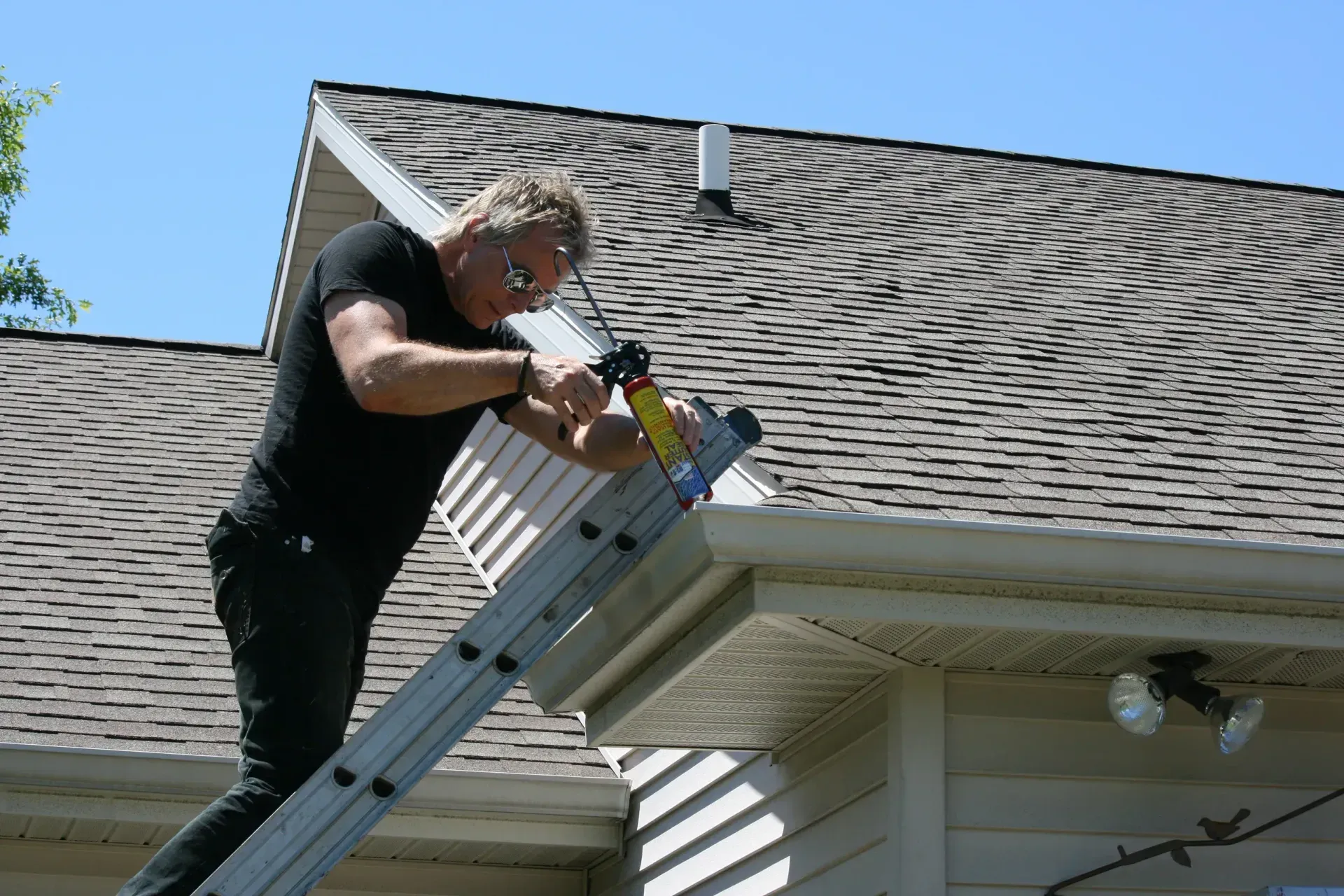 Man on ladder, applying sealant to house gutter under blue sky.