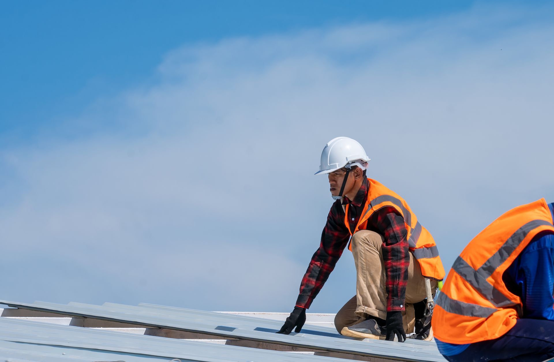 Construction workers in safety vests and hard hats on a rooftop. One kneels, inspecting the surface. Blue sky.