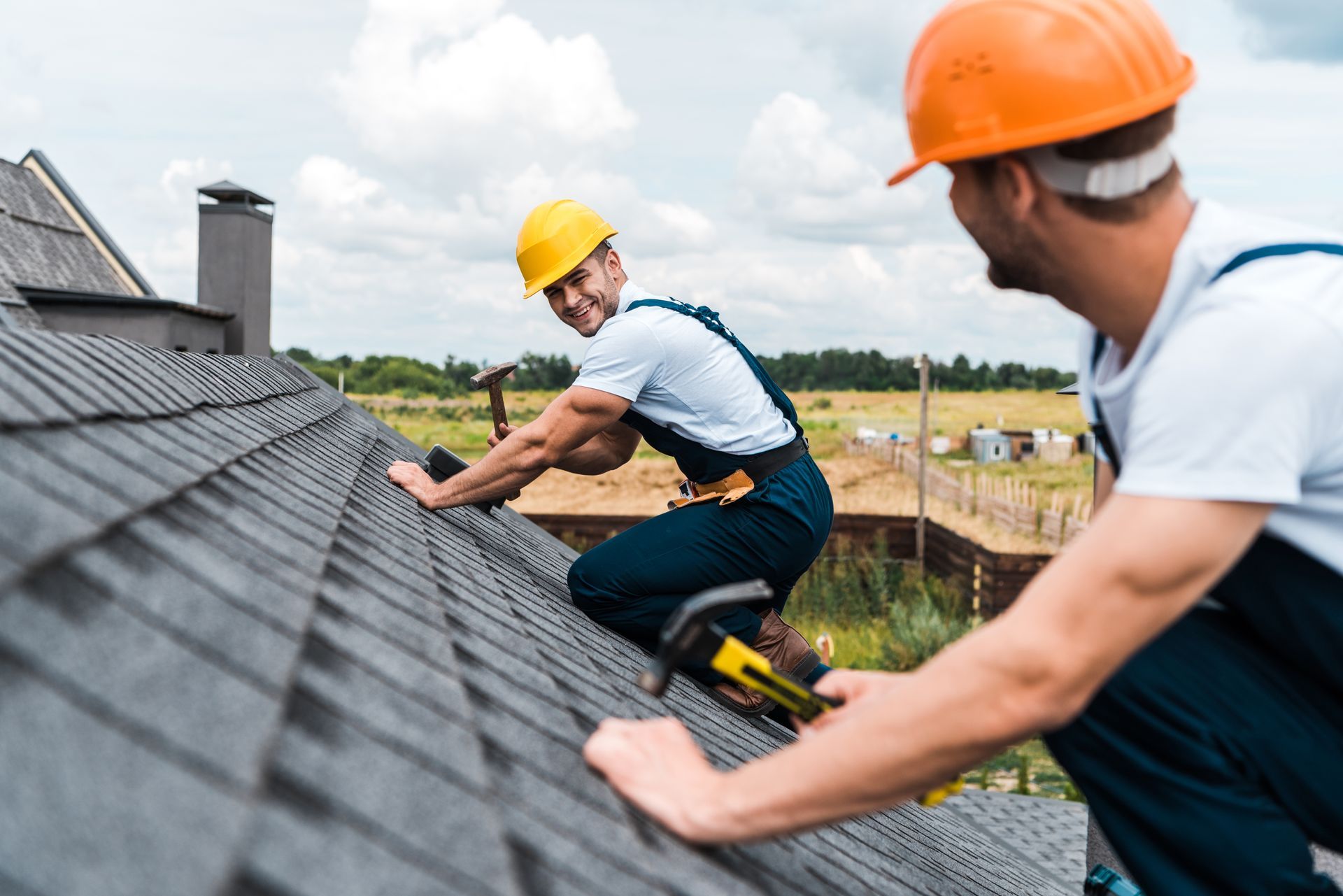 Two roofers in hard hats working on a roof, one smiles, one uses a tool.