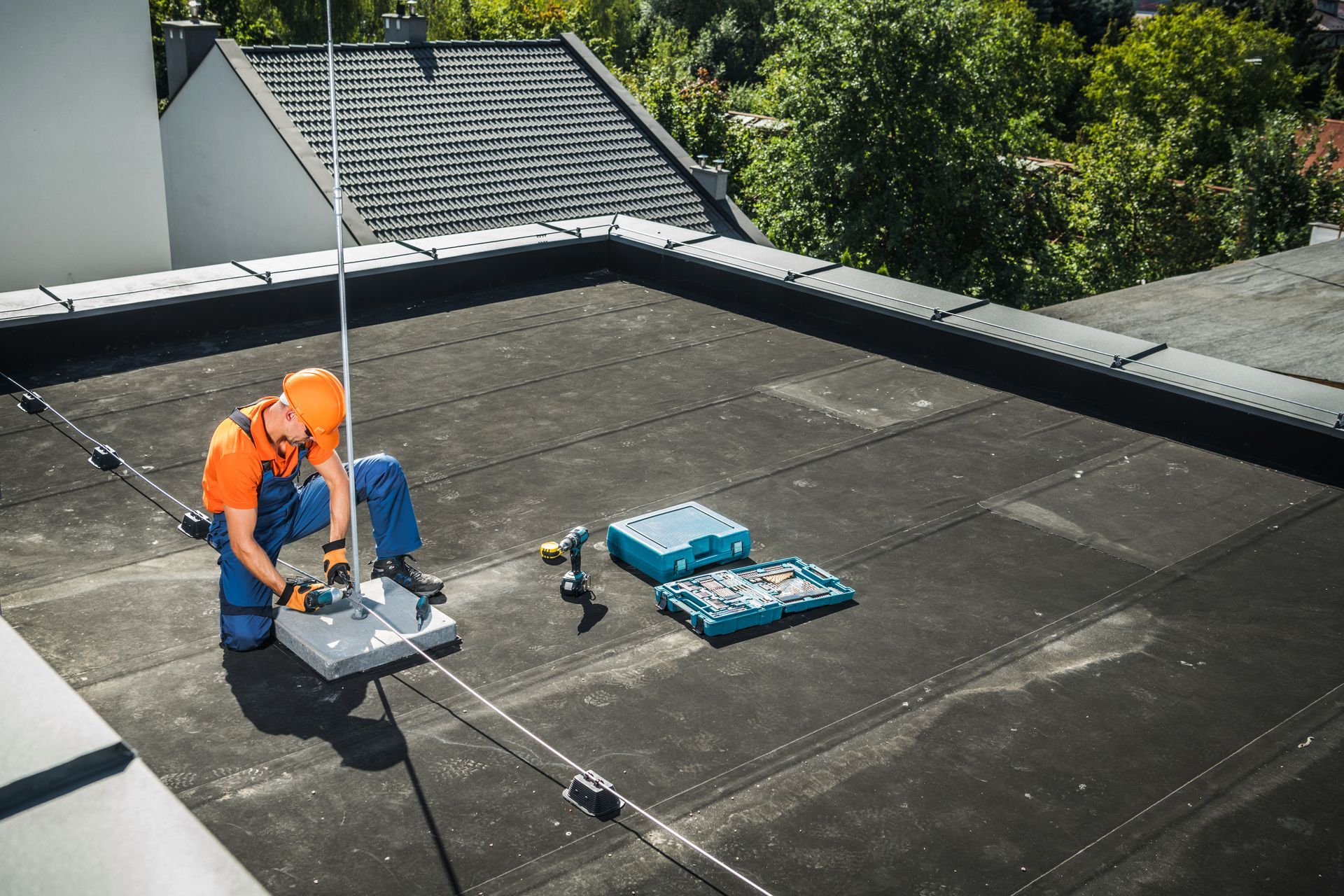 Rooftop worker in orange helmet and jumpsuit installing a lightning rod. Tools and a toolbox sit nearby.