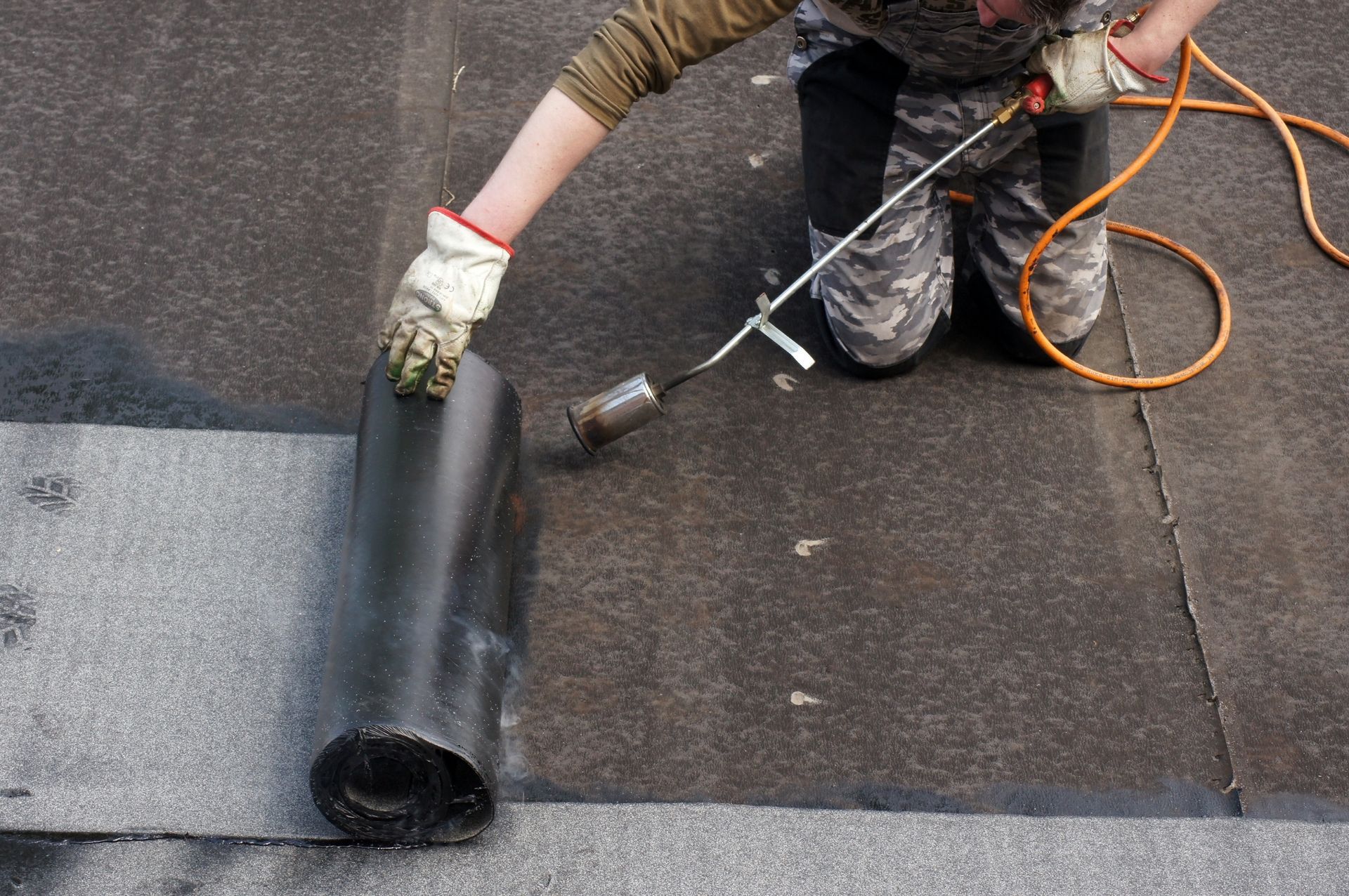 Person kneeling on a flat roof, using a torch to apply rolled roofing material.