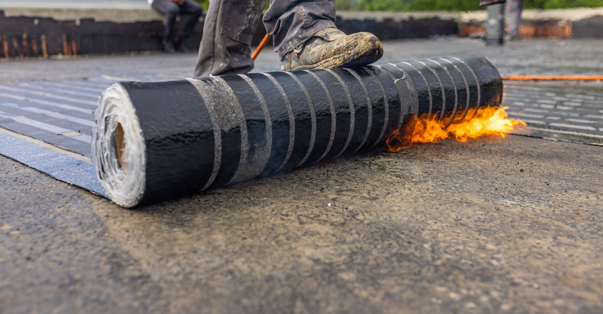 Roofer using a torch to install roofing material on a flat roof; flames visible, close-up shot.