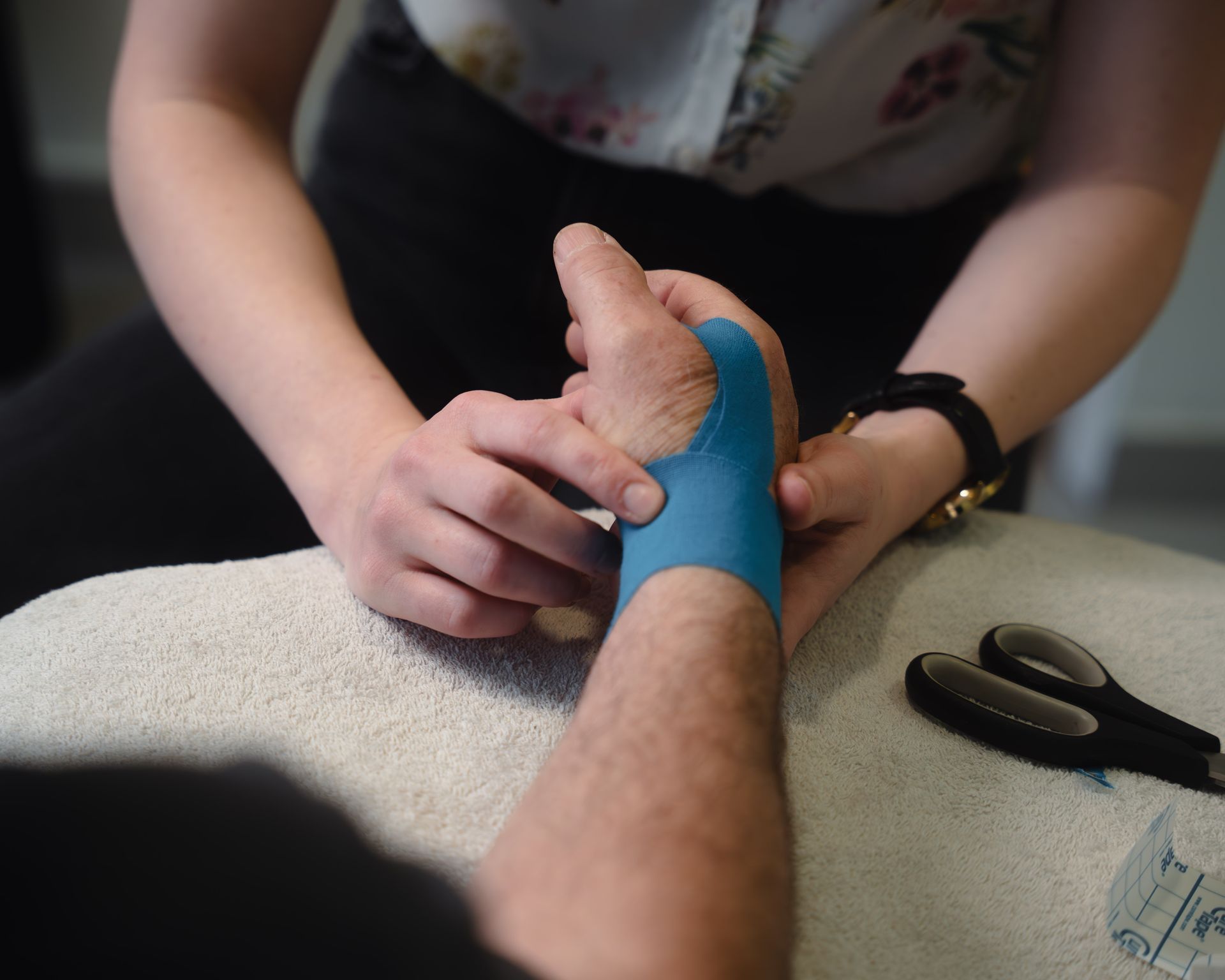 De pols van een persoon met blauwe sporttape die door iemand anders wordt aangebracht, naast een schaar op een handdoek.