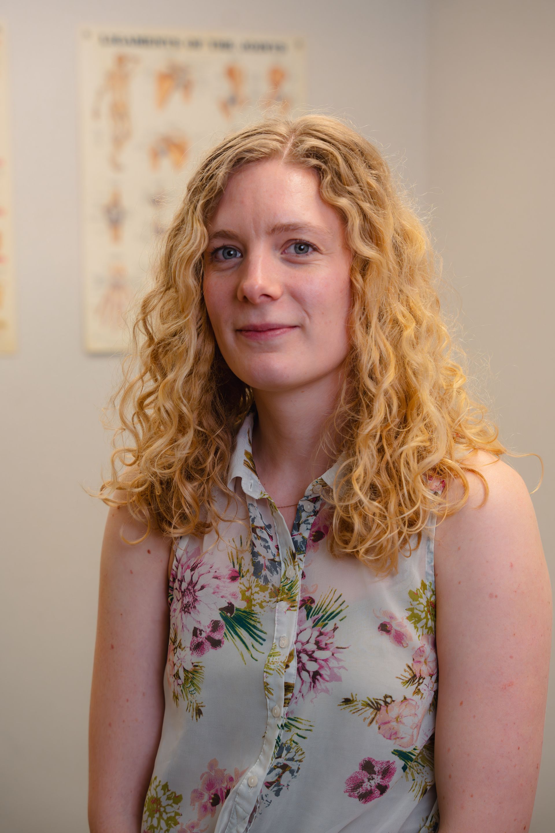 Vrouw met krullend blond haar in een topje met bloemenprint, staand in kantooromgeving, kijkend naar de camera.
