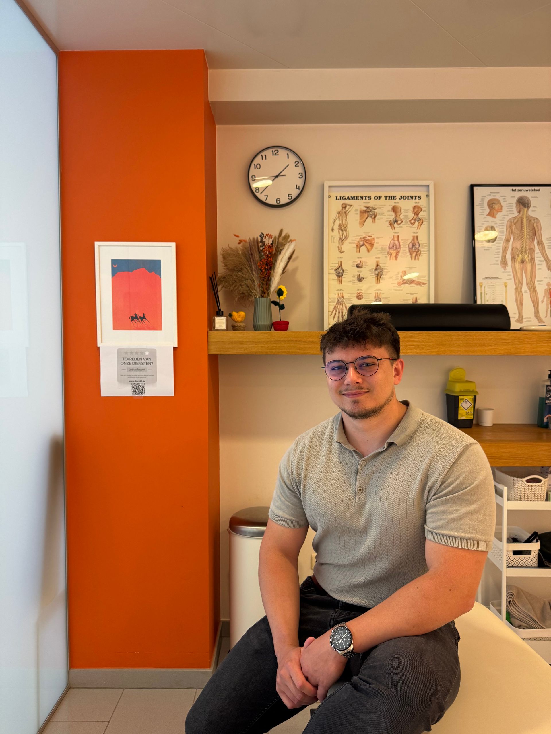 Man met bril zit in een dokterspraktijk, met een oranje muur achter zich.