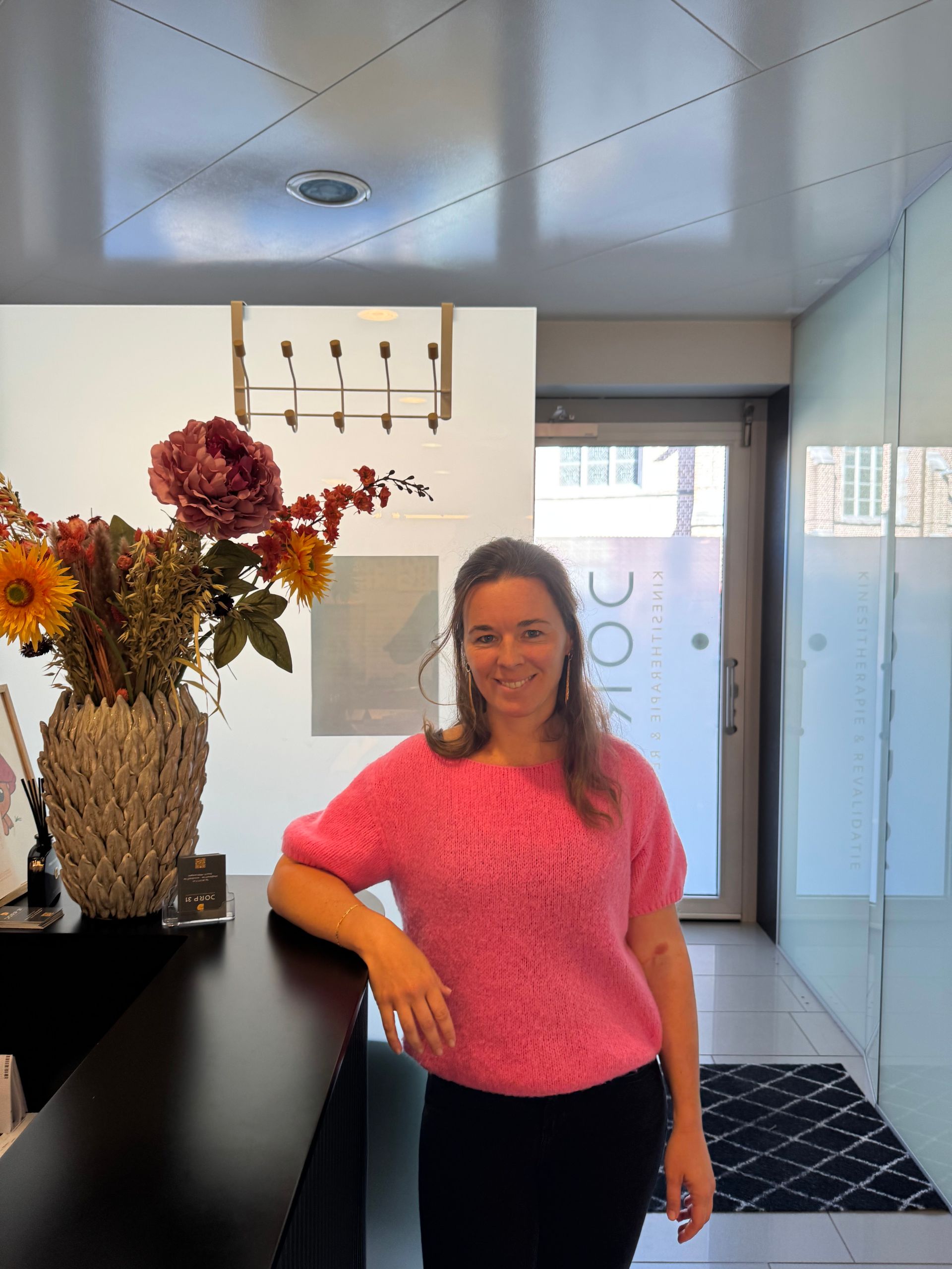 therapeute in roze trui bij een receptiebalie met bloemen.