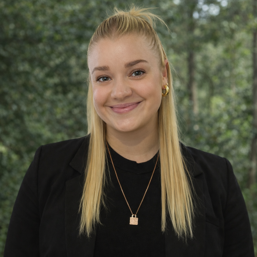 Woman wearing glasses and a necklace, smiling at the camera. She's against a plain background.
