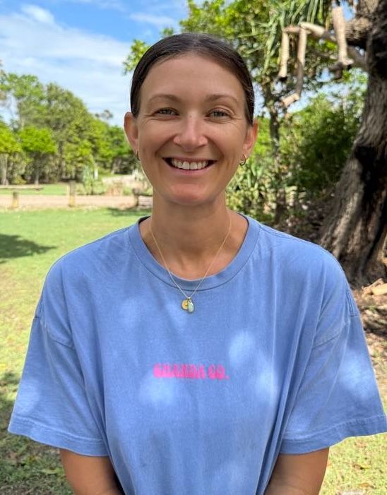 Woman smiling, wearing a blue t-shirt with a pink logo, outdoors with trees and grass in the background.