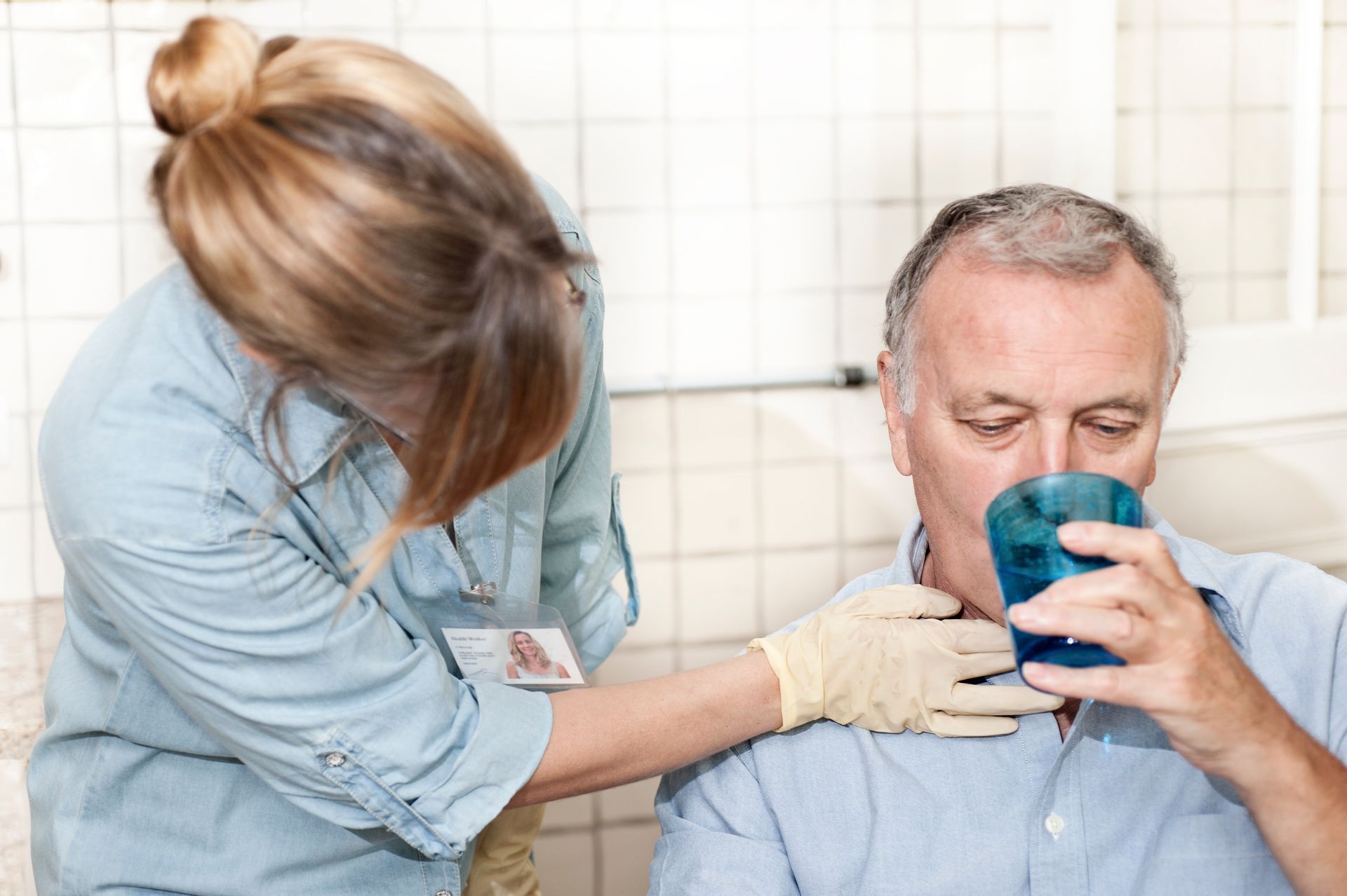 Caregiver assisting a person drinking water; gloved hand on throat, blue glass.