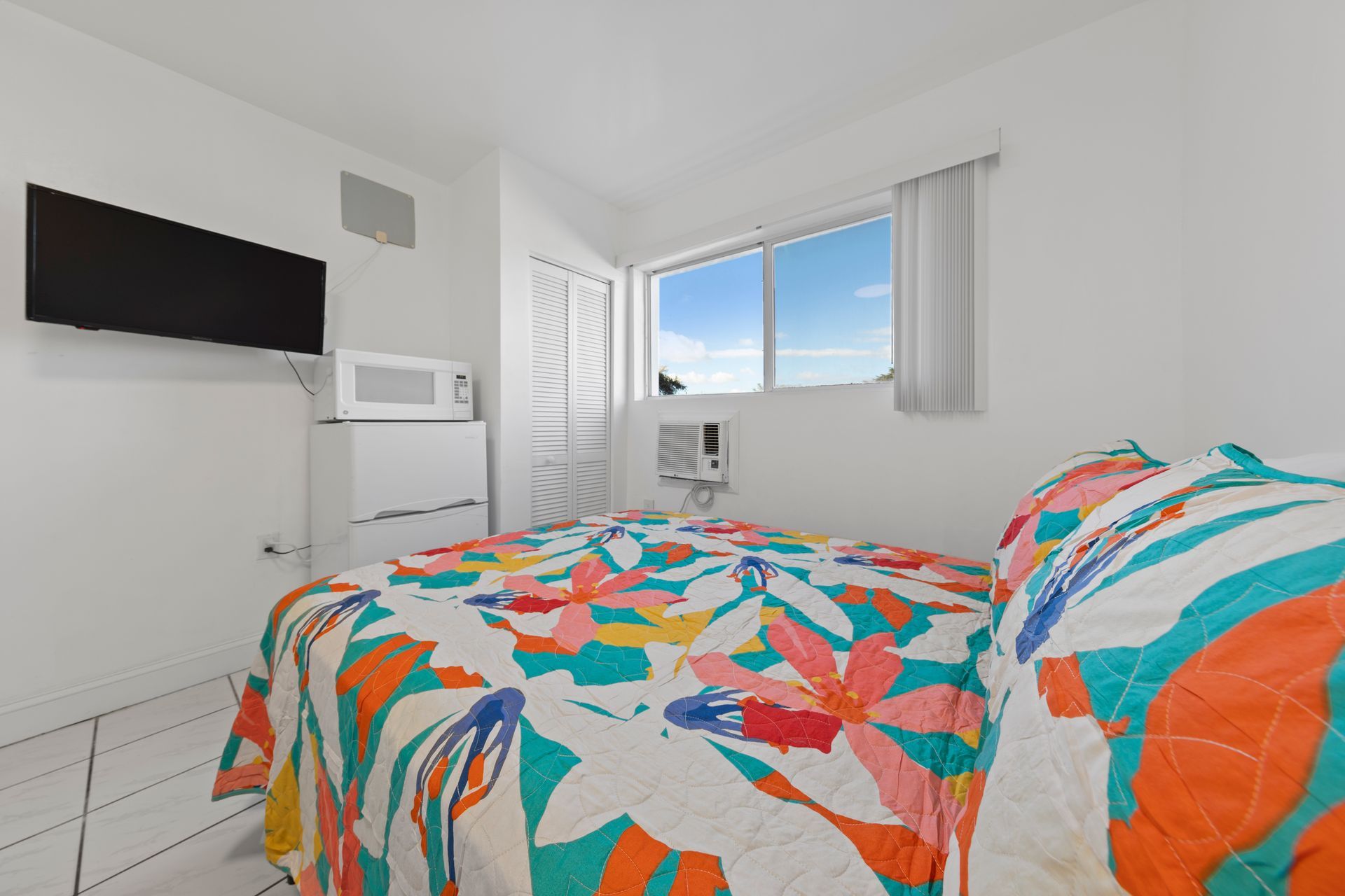 Bedroom with double bed, floral quilt, TV, microwave, window, and white walls.