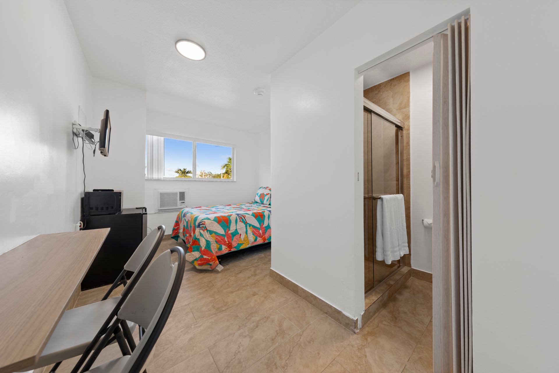 Small hotel room: bed, desk, bathroom. White walls, tile floor, bright window view.
