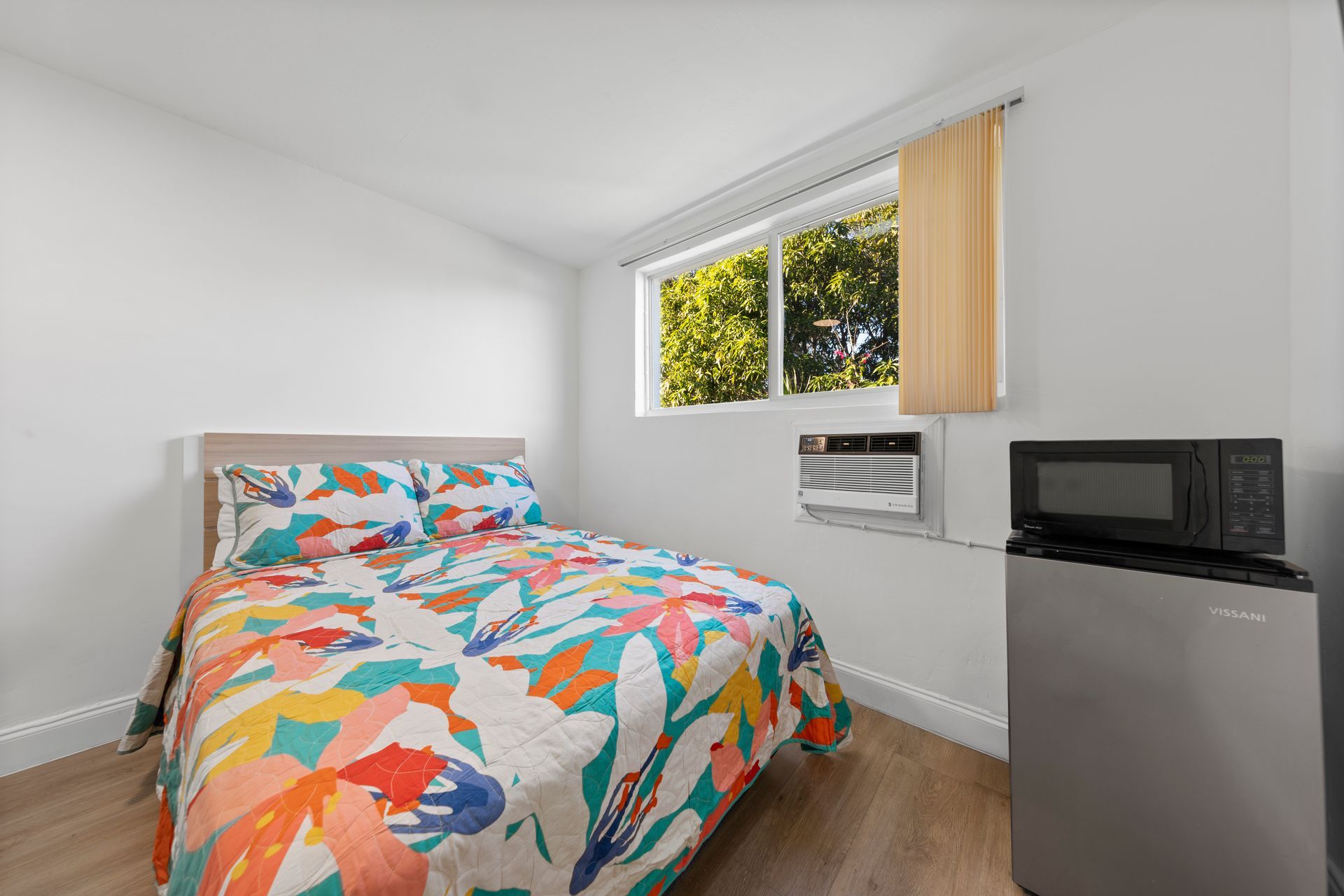 Small, simple bedroom with bed, colorful comforter, window, microwave, and mini-fridge.