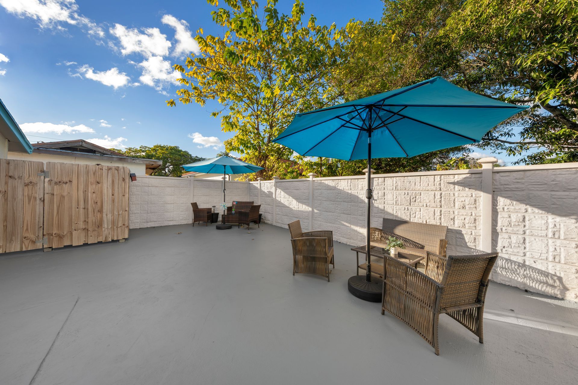 Patio with blue umbrellas, wicker furniture, and a gray floor against a white and wood fence.