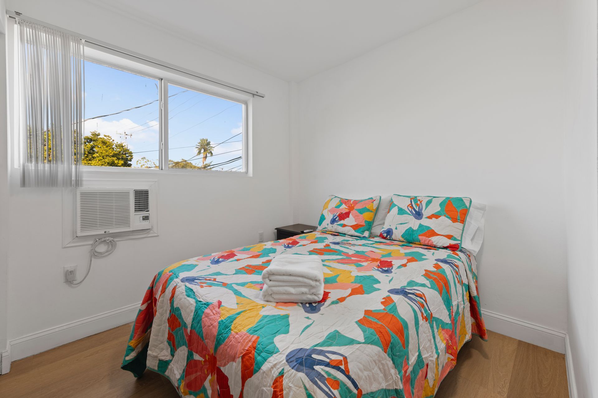 Bedroom with bed, colorful floral quilt, window with view, air conditioner.