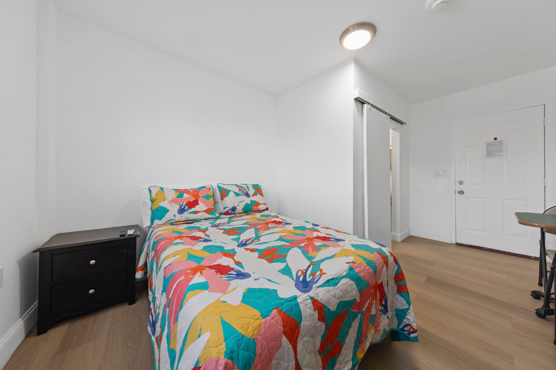 Bedroom with white walls, wooden floor, double bed with colorful floral quilt, and black nightstand.