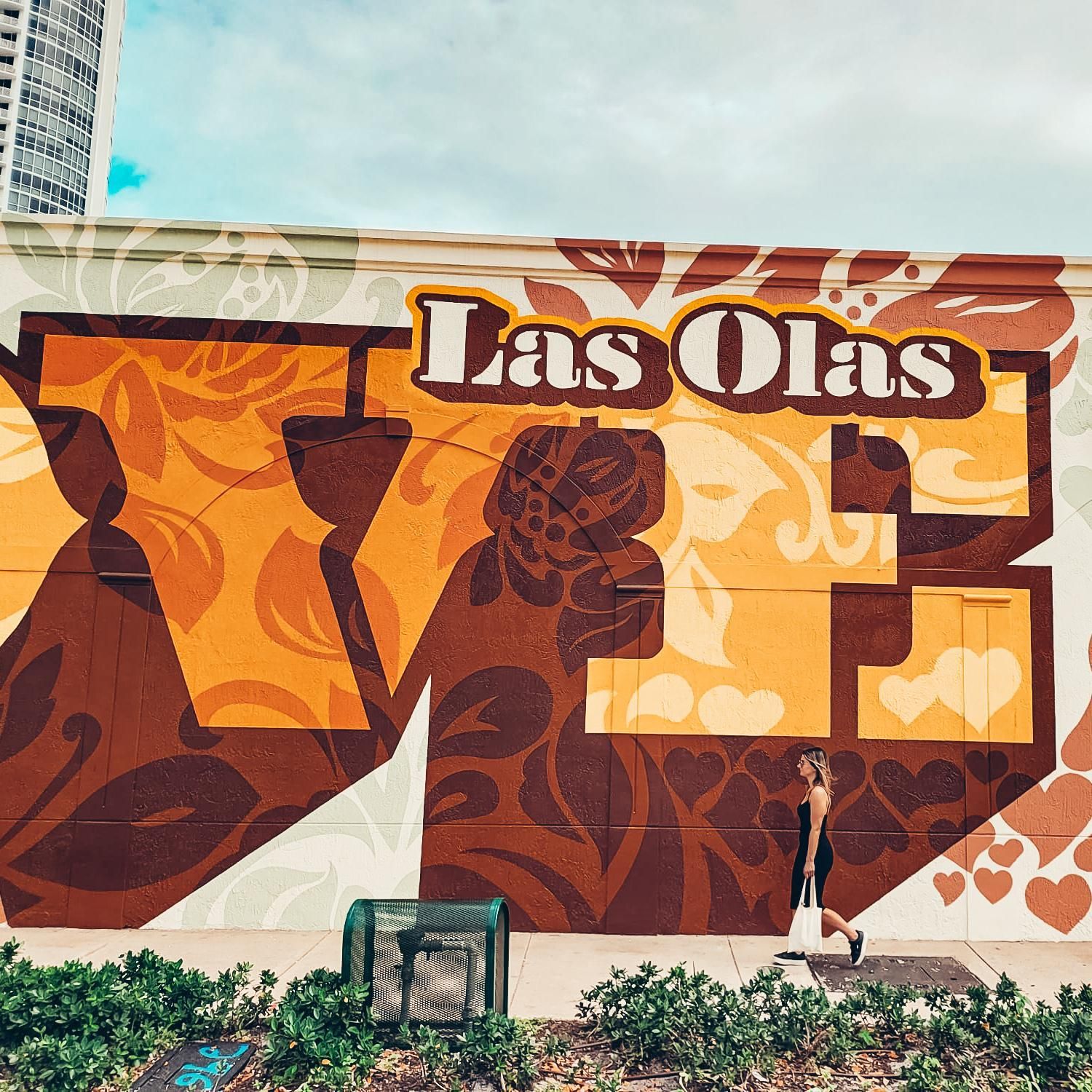 Das Wandgemälde von Las Olas zeigt eine vorbeigehende Frau vor blauem Himmel und grünen Pflanzen.