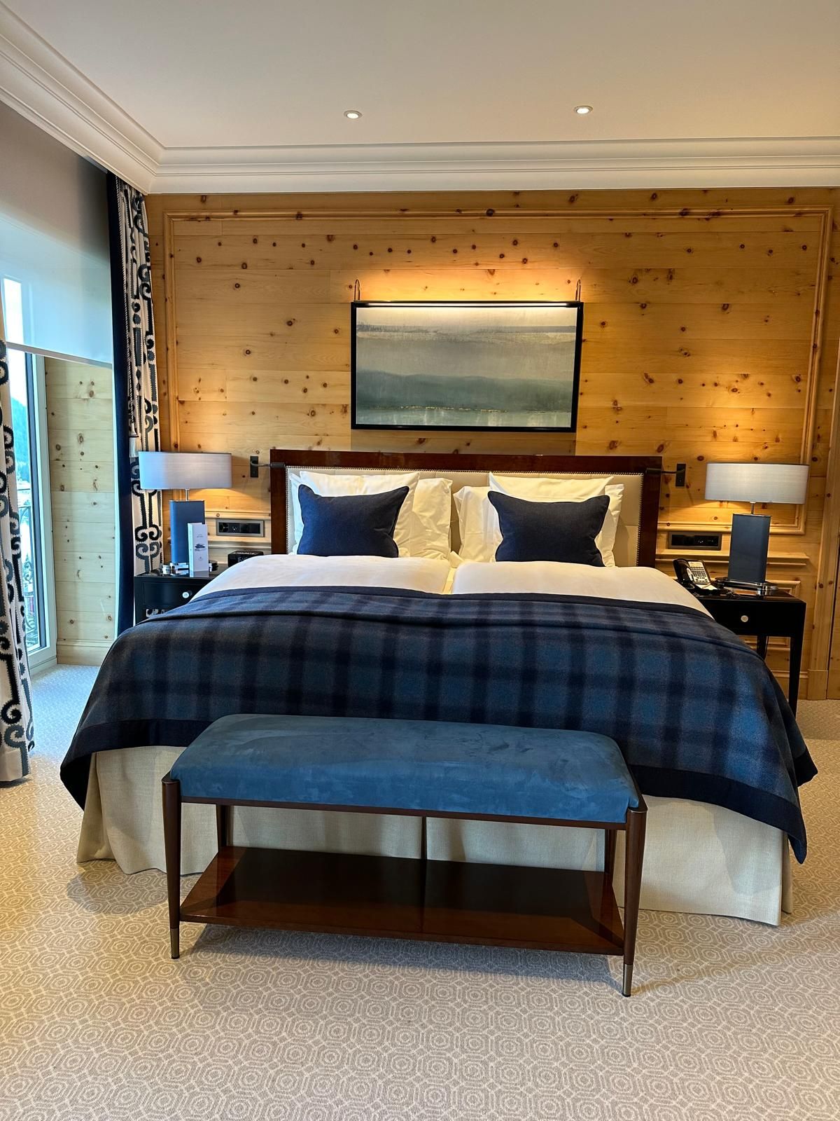 Schlafzimmer mit holzgetäfelter Wand, Queensize-Bett mit blau-karierter Decke, blauer Bank und zwei Nachttischen.