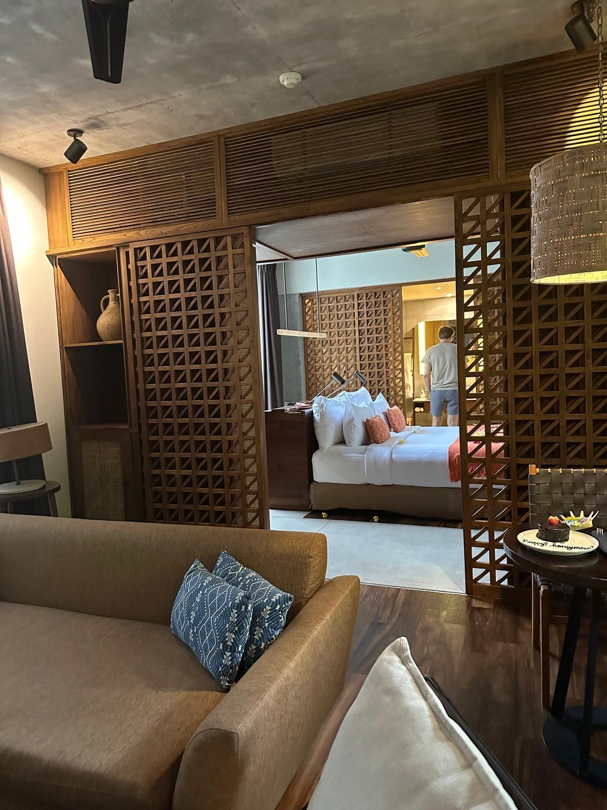 Eine Hotelsuite mit einem hellbraunen Sofa, hölzernen Raumteilern und Blick auf ein Bett mit weissen und orangefarbenen Kissen.