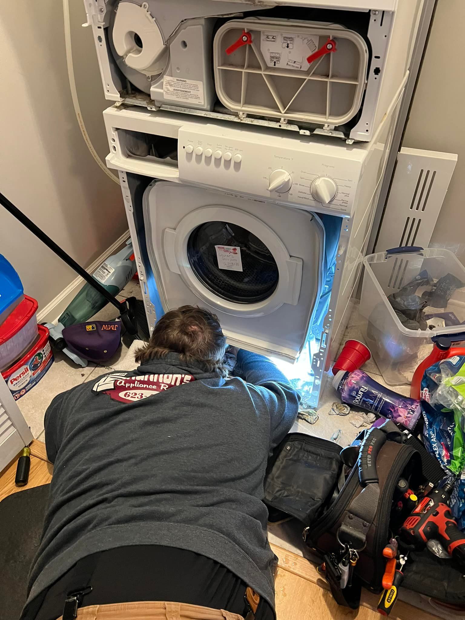 A man is kneeling down in front of a stacked washer and dryer.