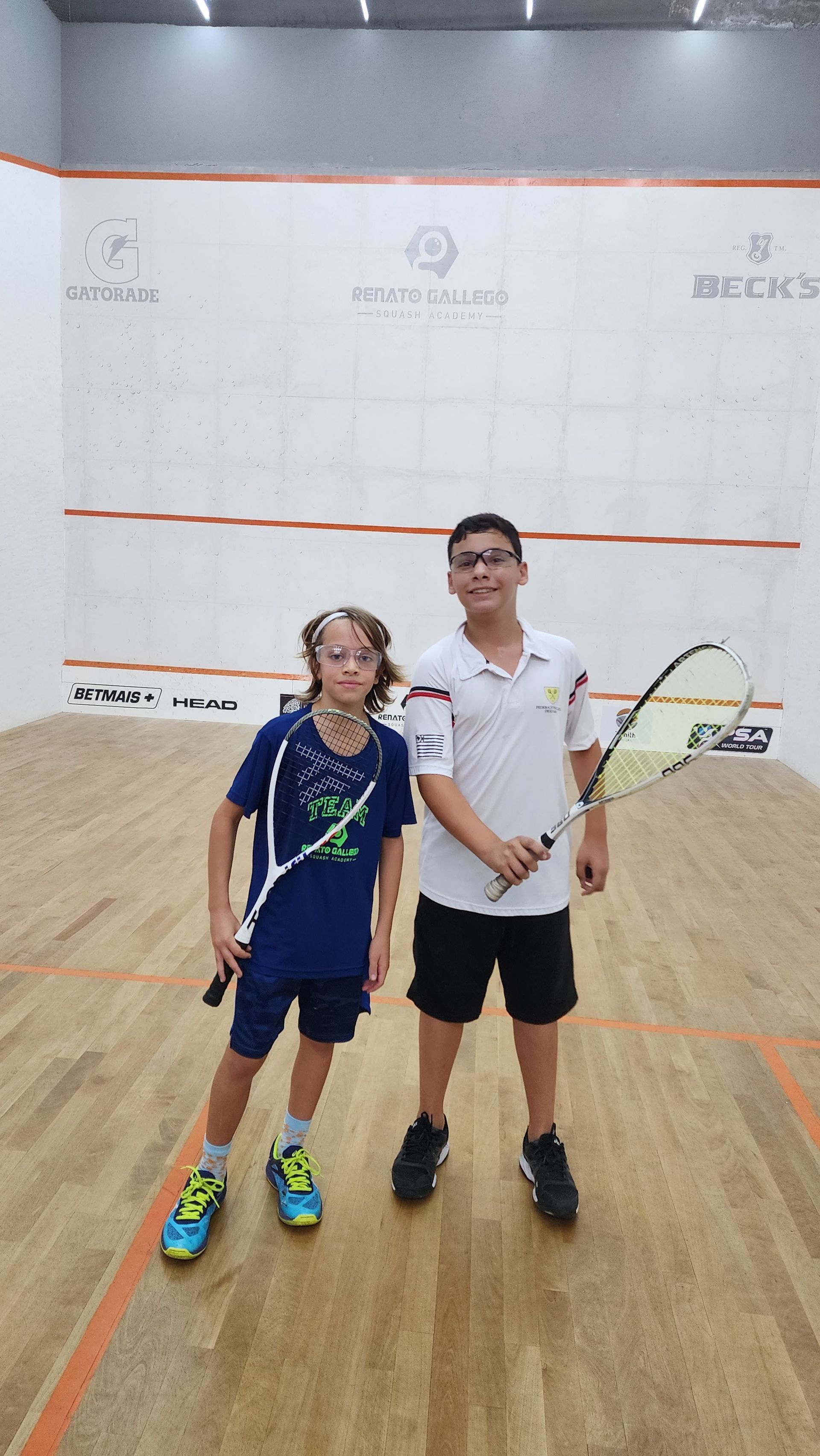Dois meninos estão em uma quadra de squash segurando raquetes.
